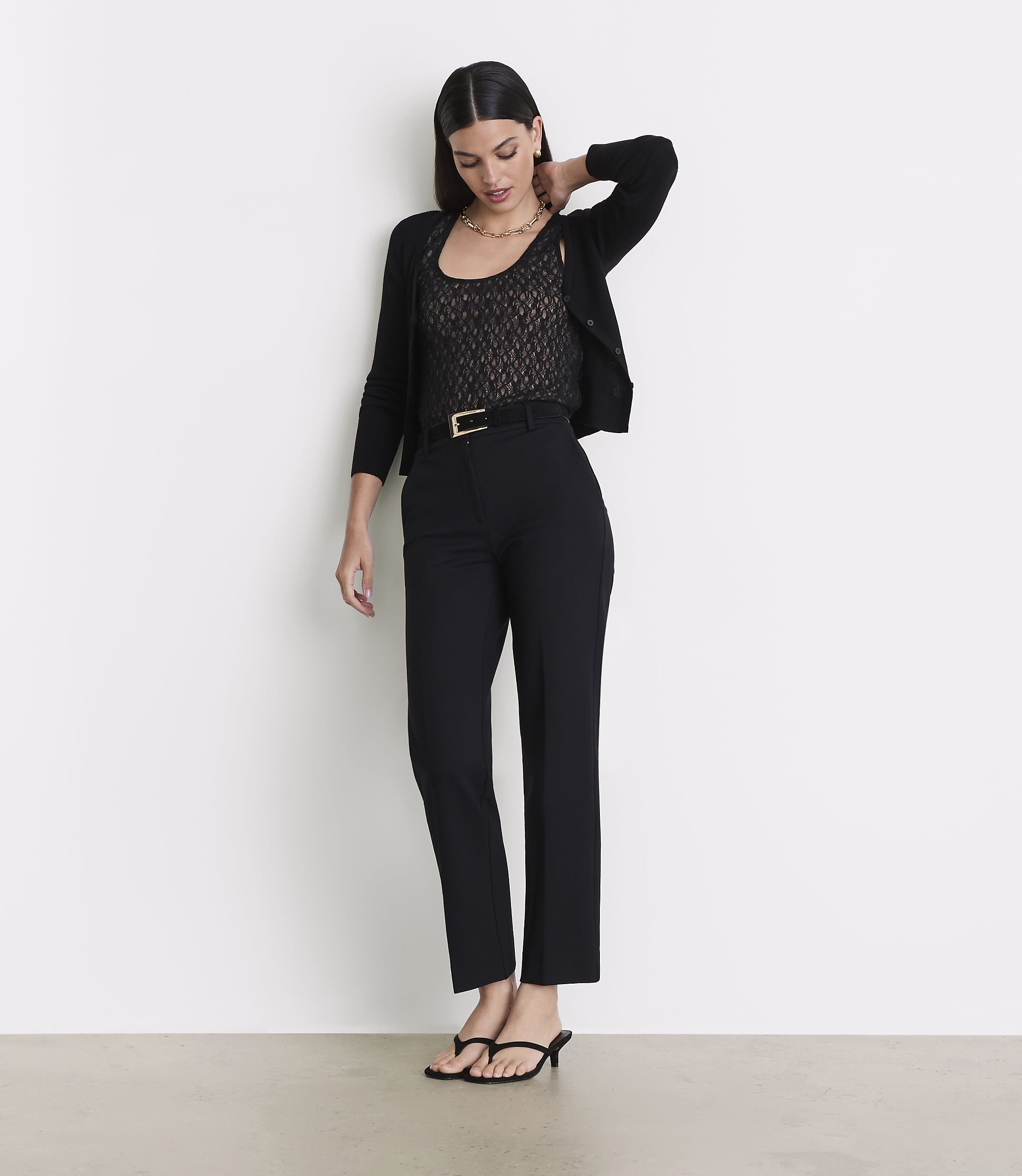 A woman in a black cardigan, lace top, and black pants stands against a plain white background.