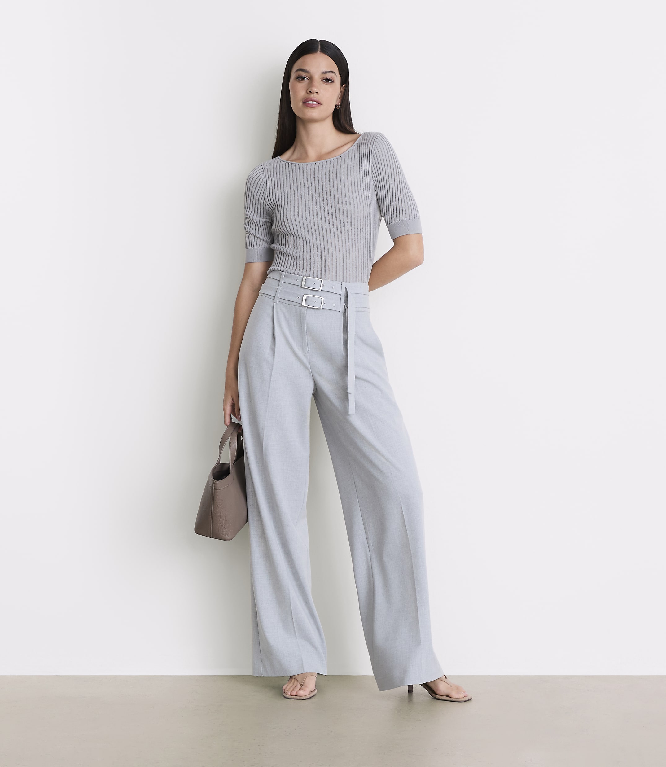 A woman in a grey ribbed top and light blue wide-leg pants stands against a plain white background, holding a taupe handbag.