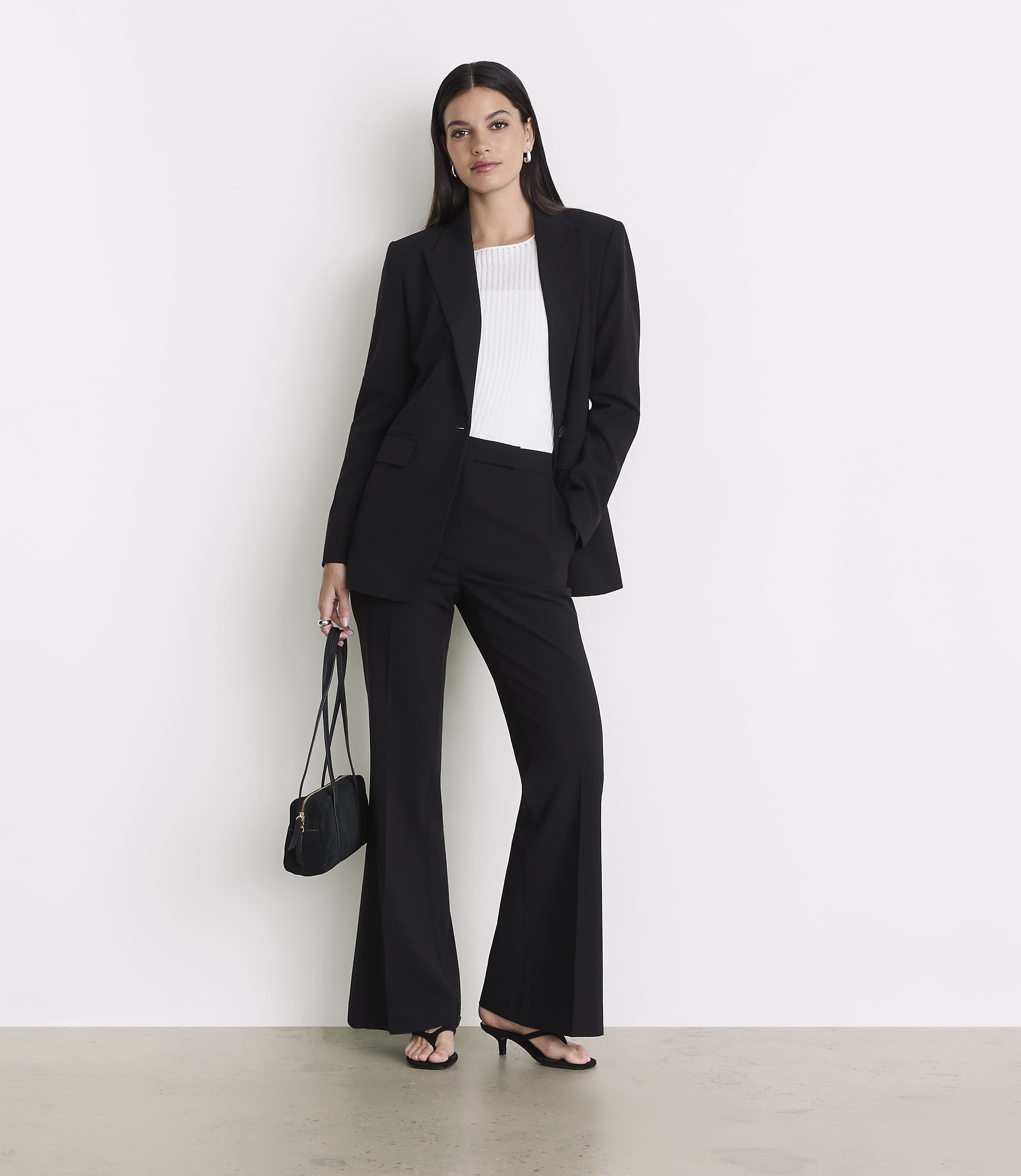 A woman in a black suit and white top stands against a white wall, holding a black purse.