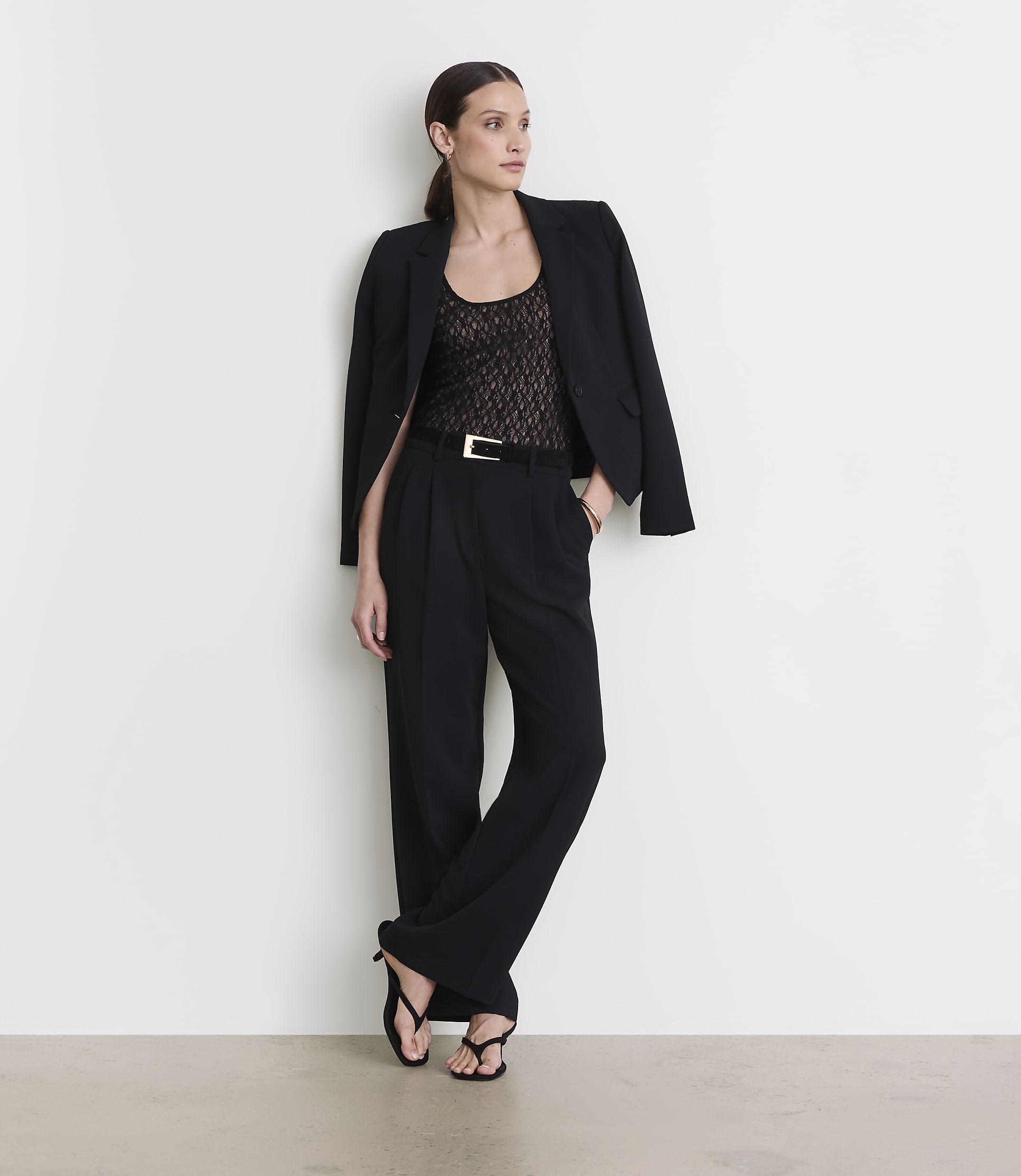 A woman in a black blazer, lace top, and wide-leg pants stands against a plain white wall.