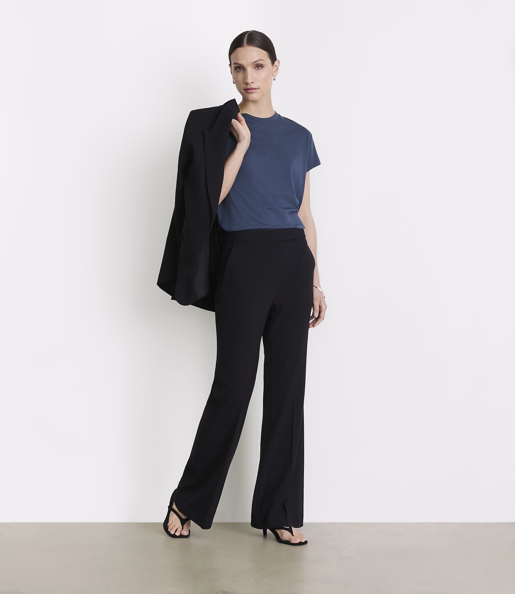 A woman in a blue t-shirt and black pants stands against a white background, with a black blazer draped over her shoulder.