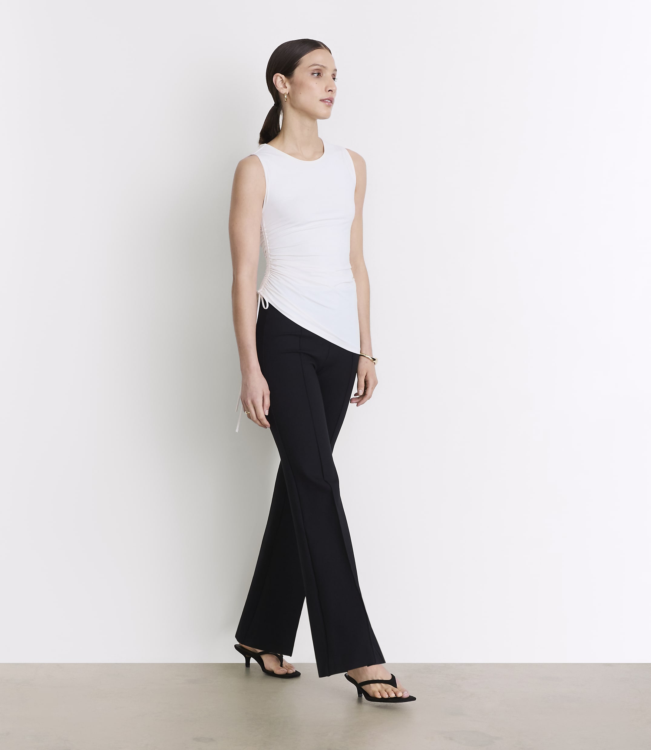 A woman in a white sleeveless top and black pants stands against a plain white background.