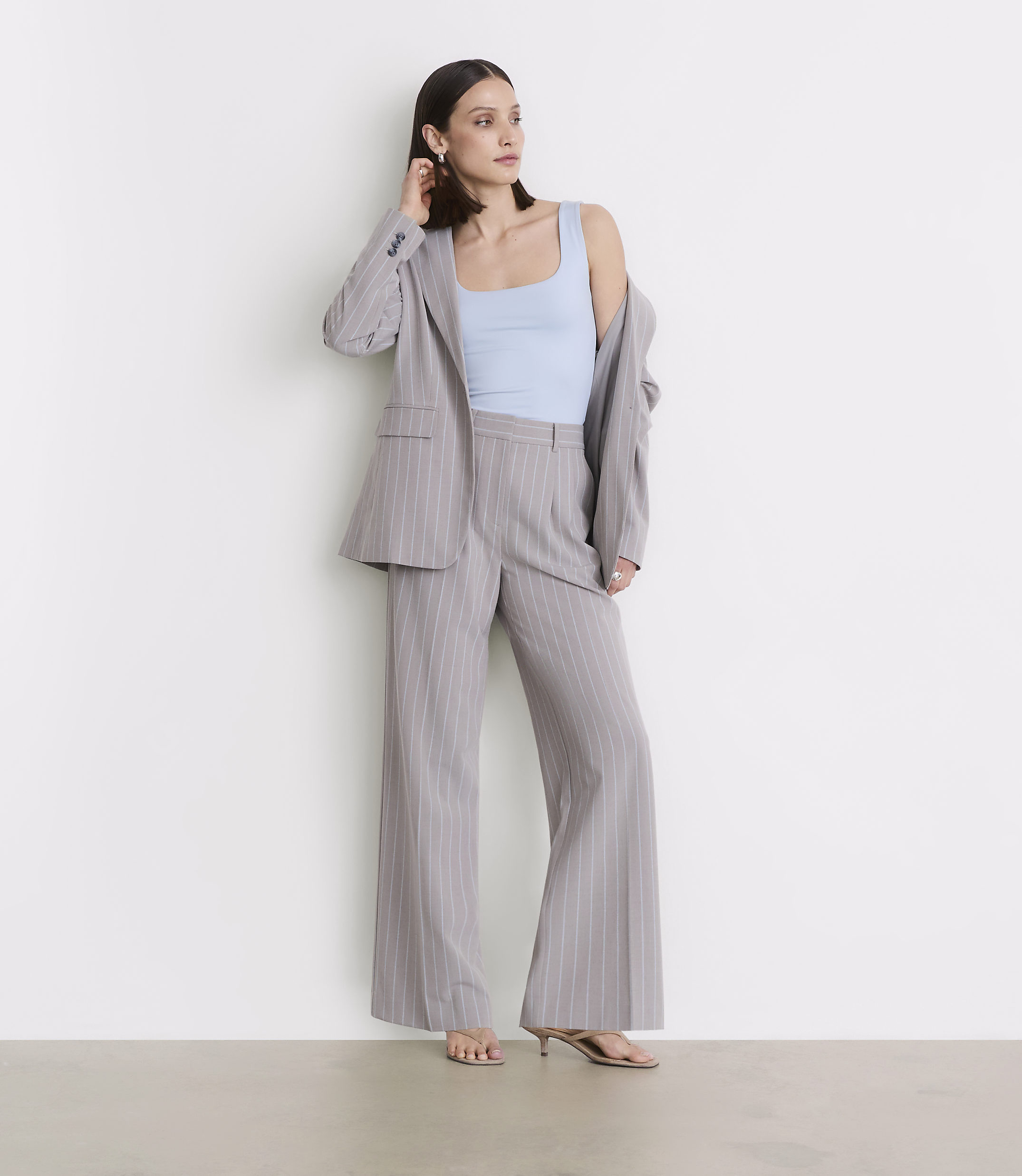A woman in a light blue tank top and a pinstripe suit stands against a plain white wall.