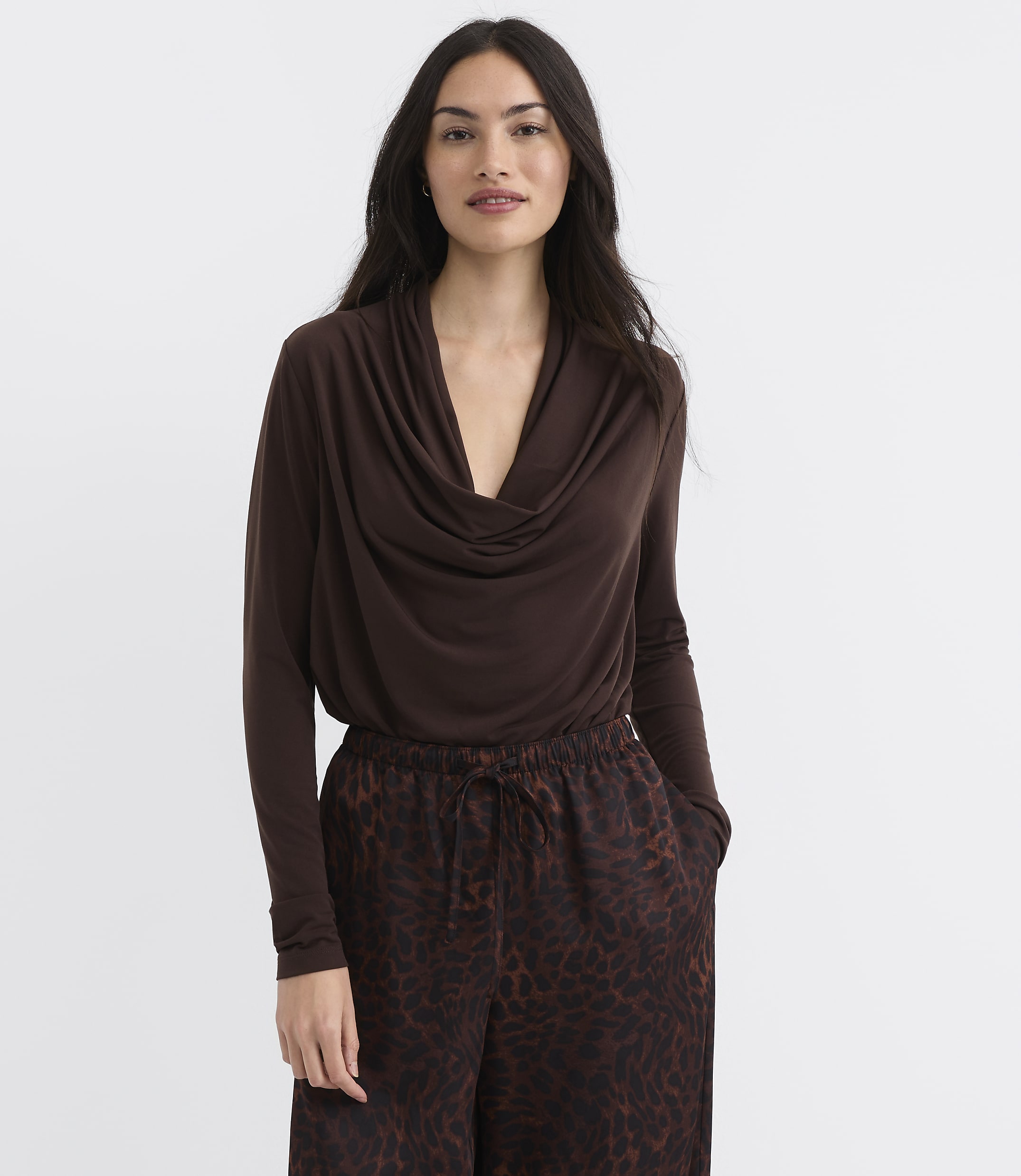A woman in a brown cowl-neck top and leopard-print pants stands against a plain white background.