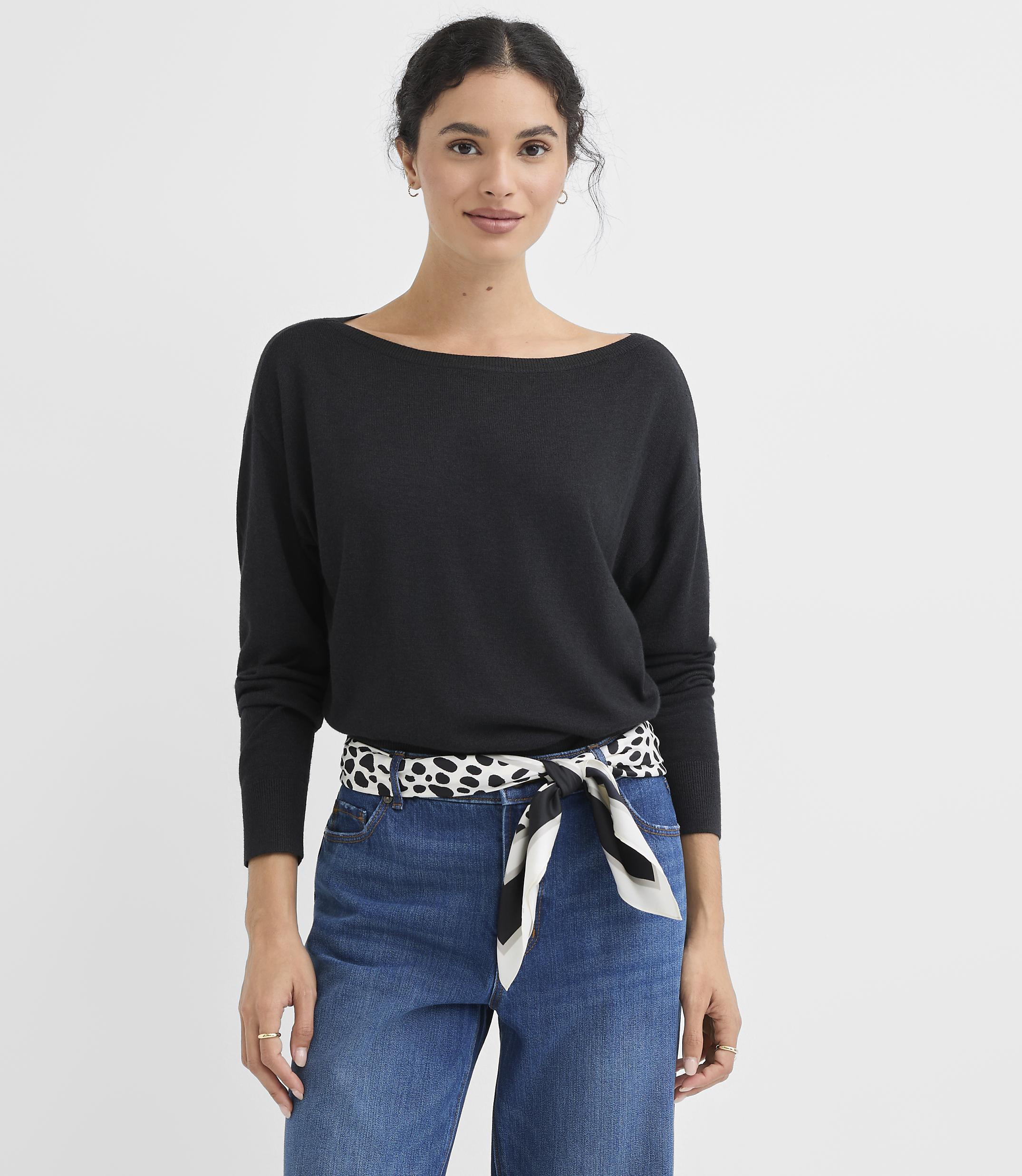 A woman in a black sweater and blue jeans stands against a plain white background.