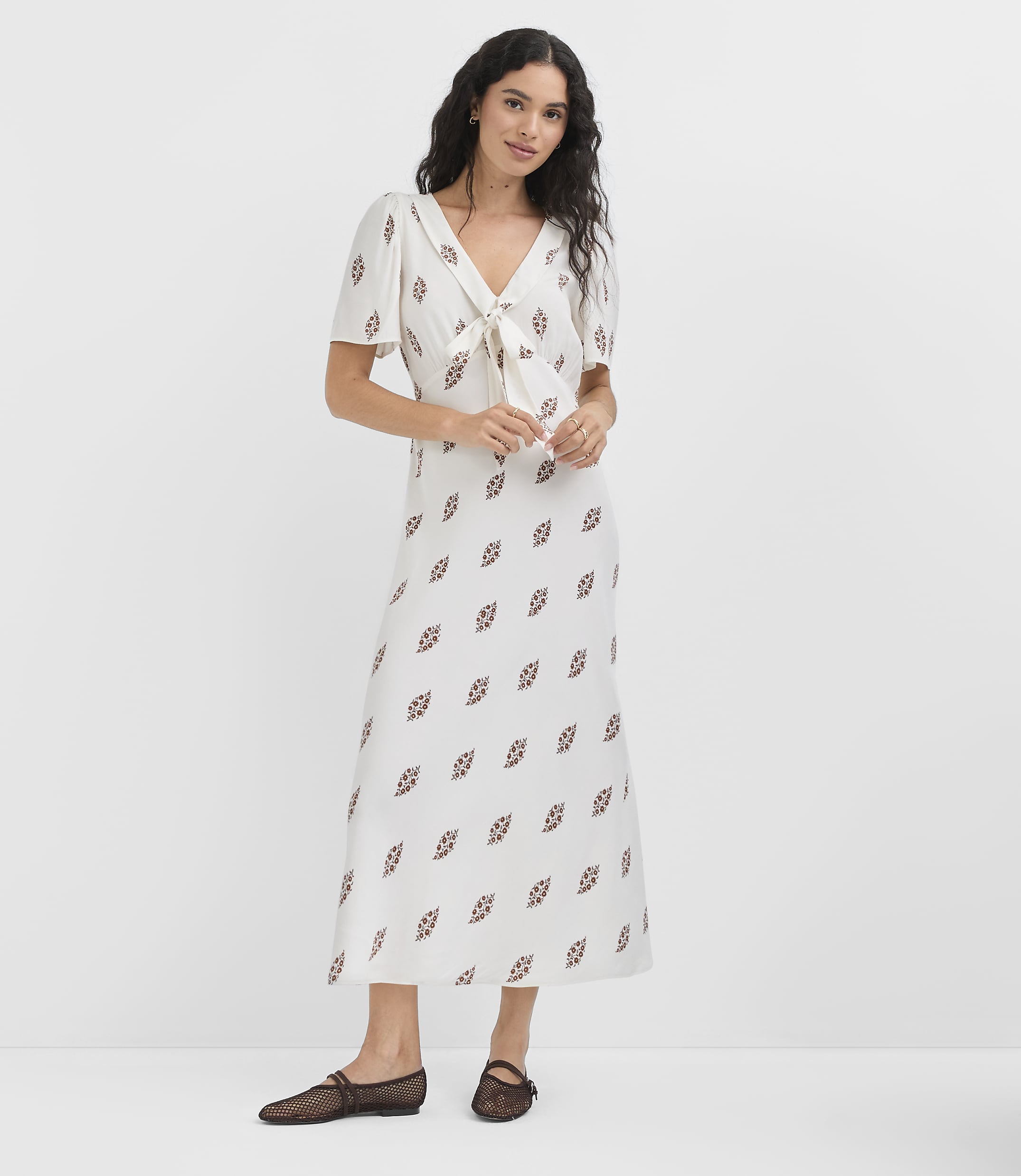 A woman in a white patterned dress and black flats stands against a plain white background.