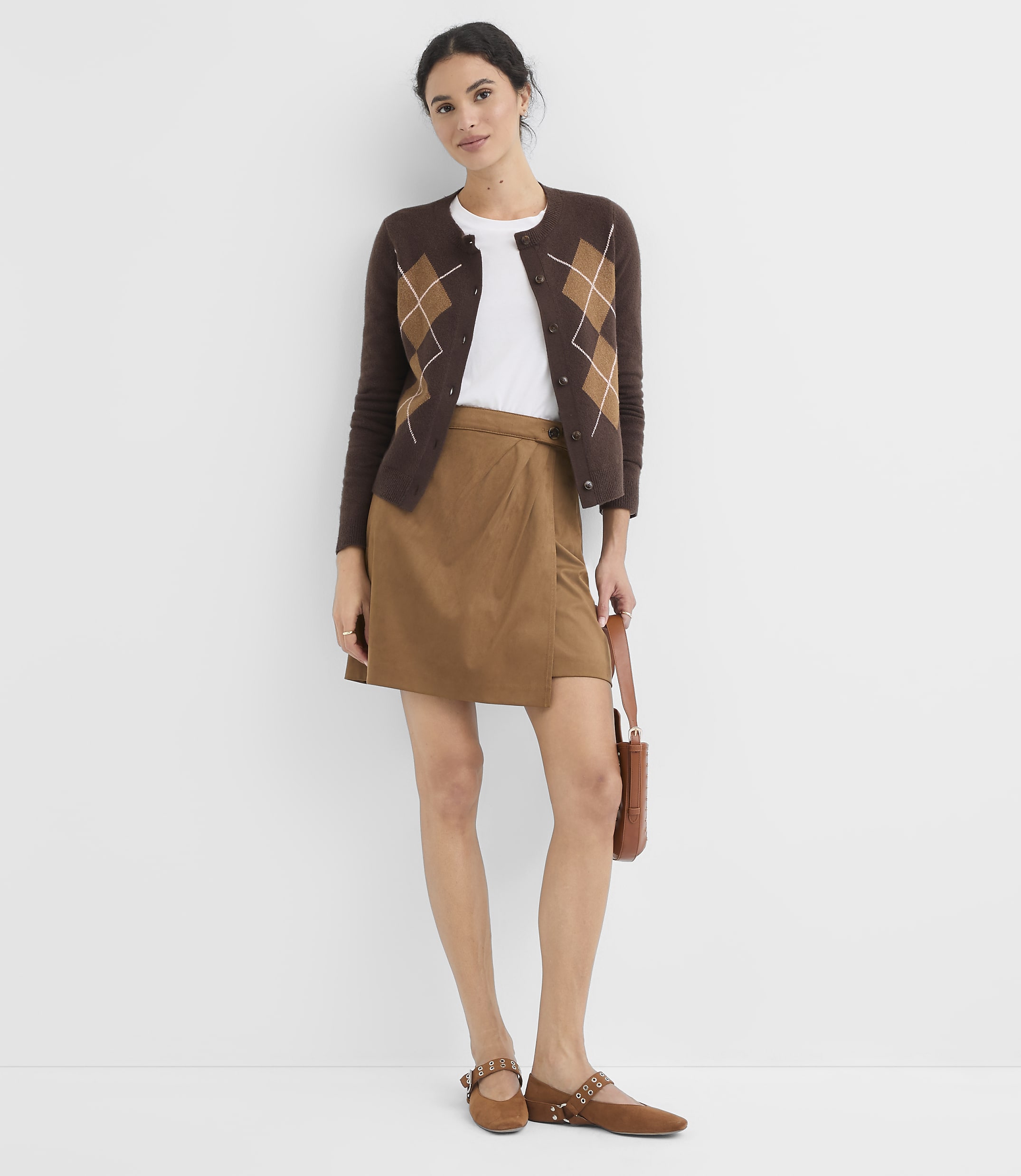 A woman in a brown argyle cardigan and tan skirt stands against a plain white background, holding a brown purse.