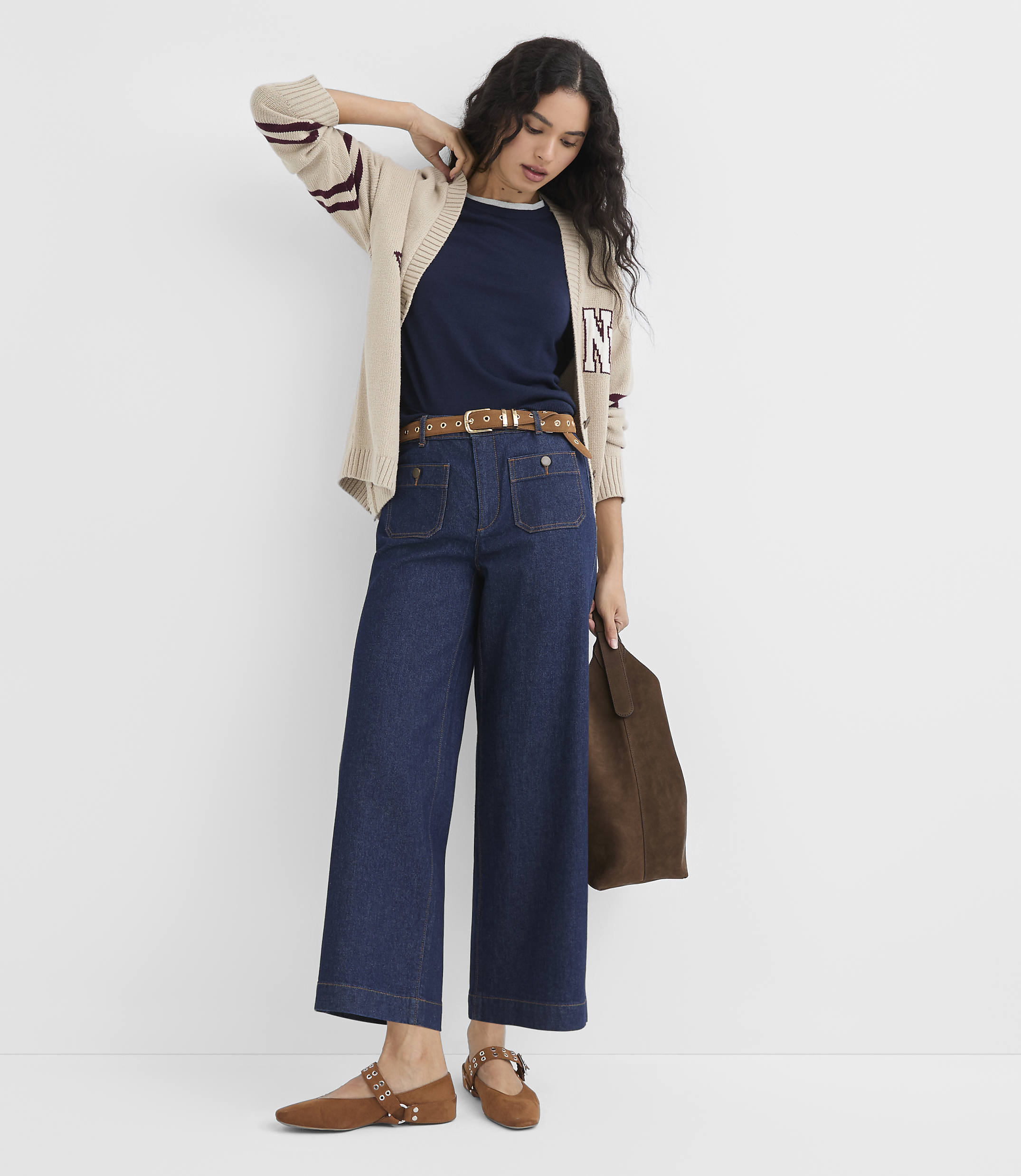 A woman in a navy shirt, beige cardigan, and wide-leg jeans stands against a plain white background, holding a brown bag.