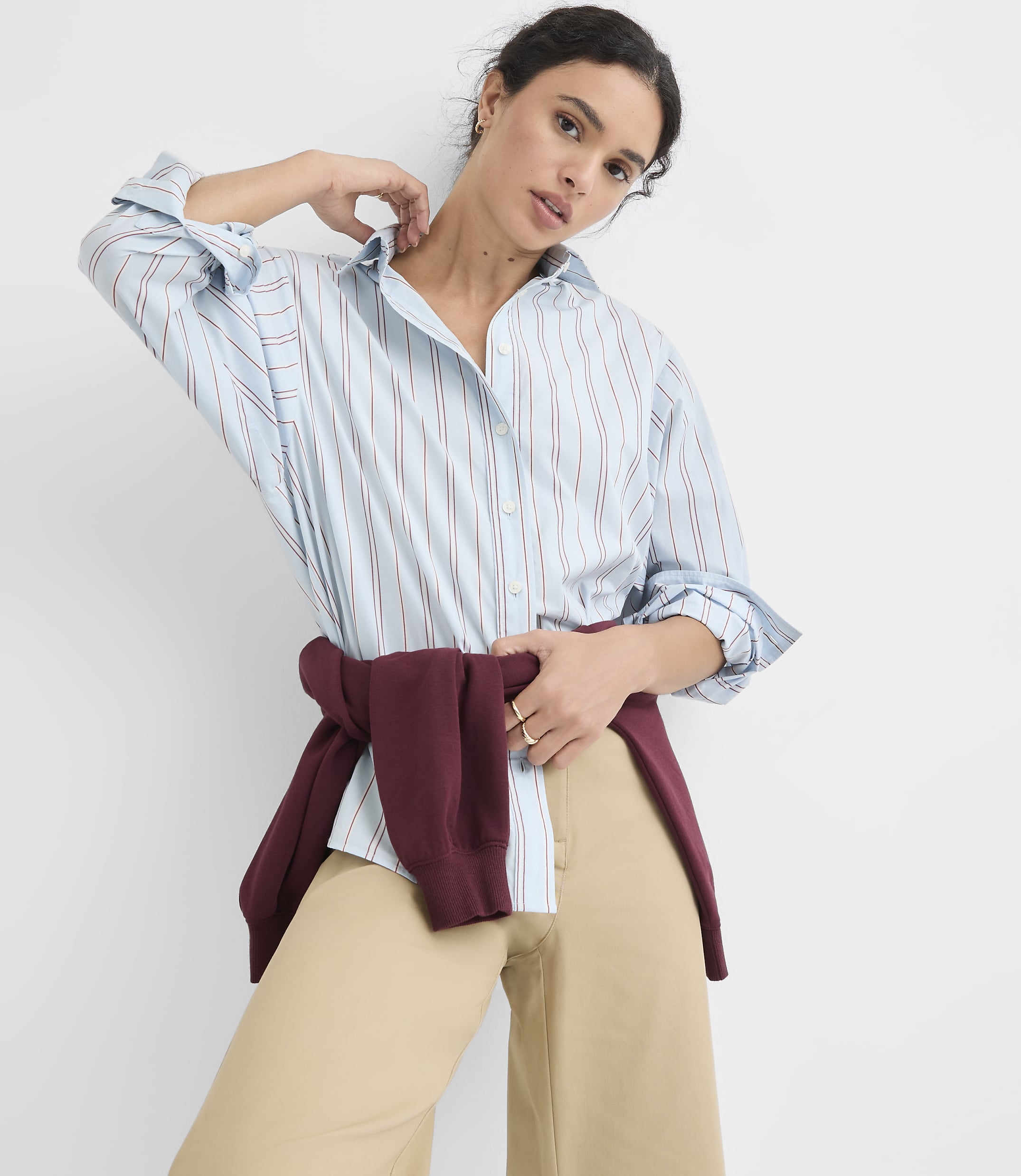A woman in a light blue pinstripe shirt and tan pants poses against a plain white background, with a maroon sweater tied around her waist.