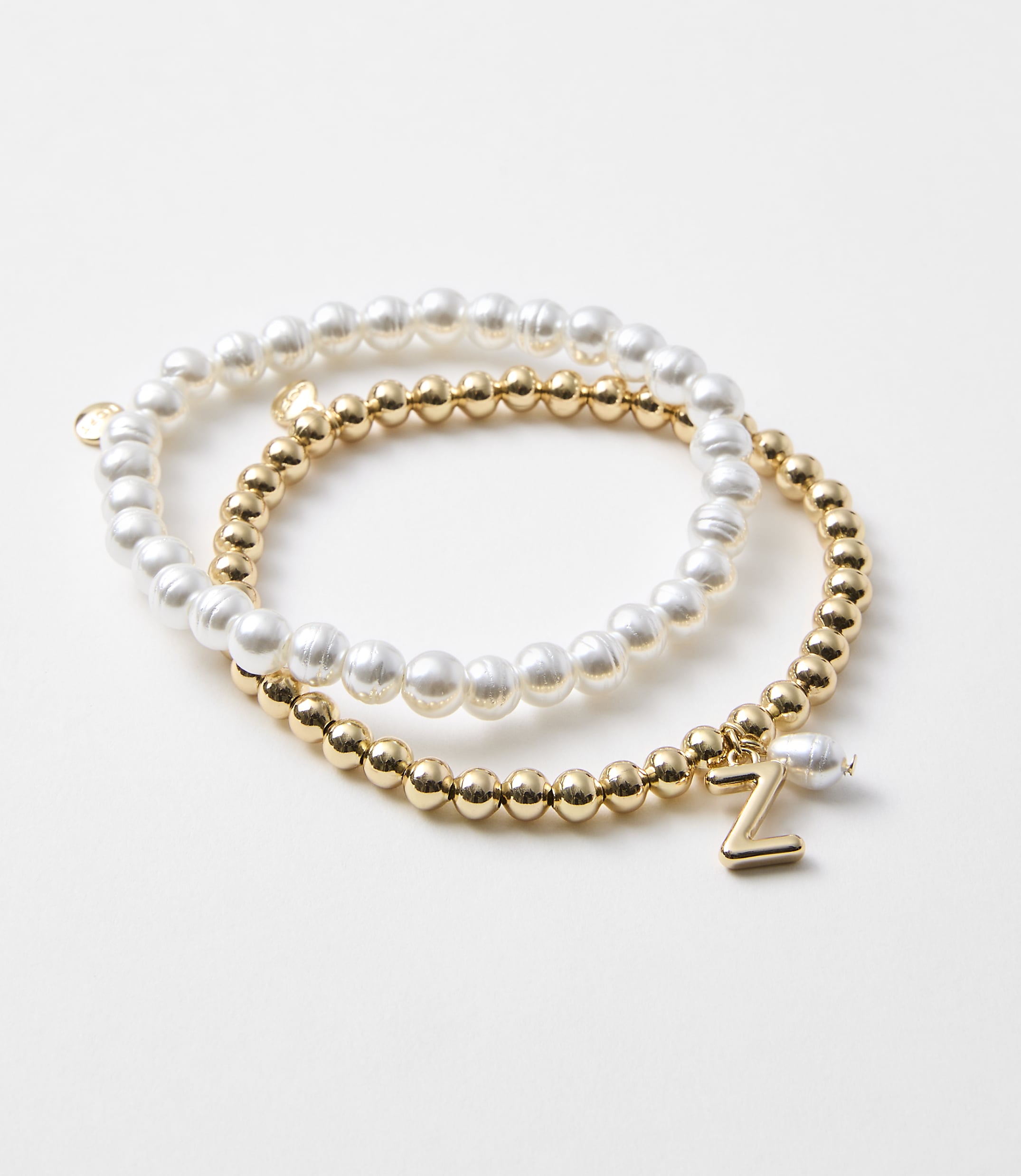 Two bracelets, one with white pearls and the other with gold beads and a "Z" charm, rest on a plain white background.