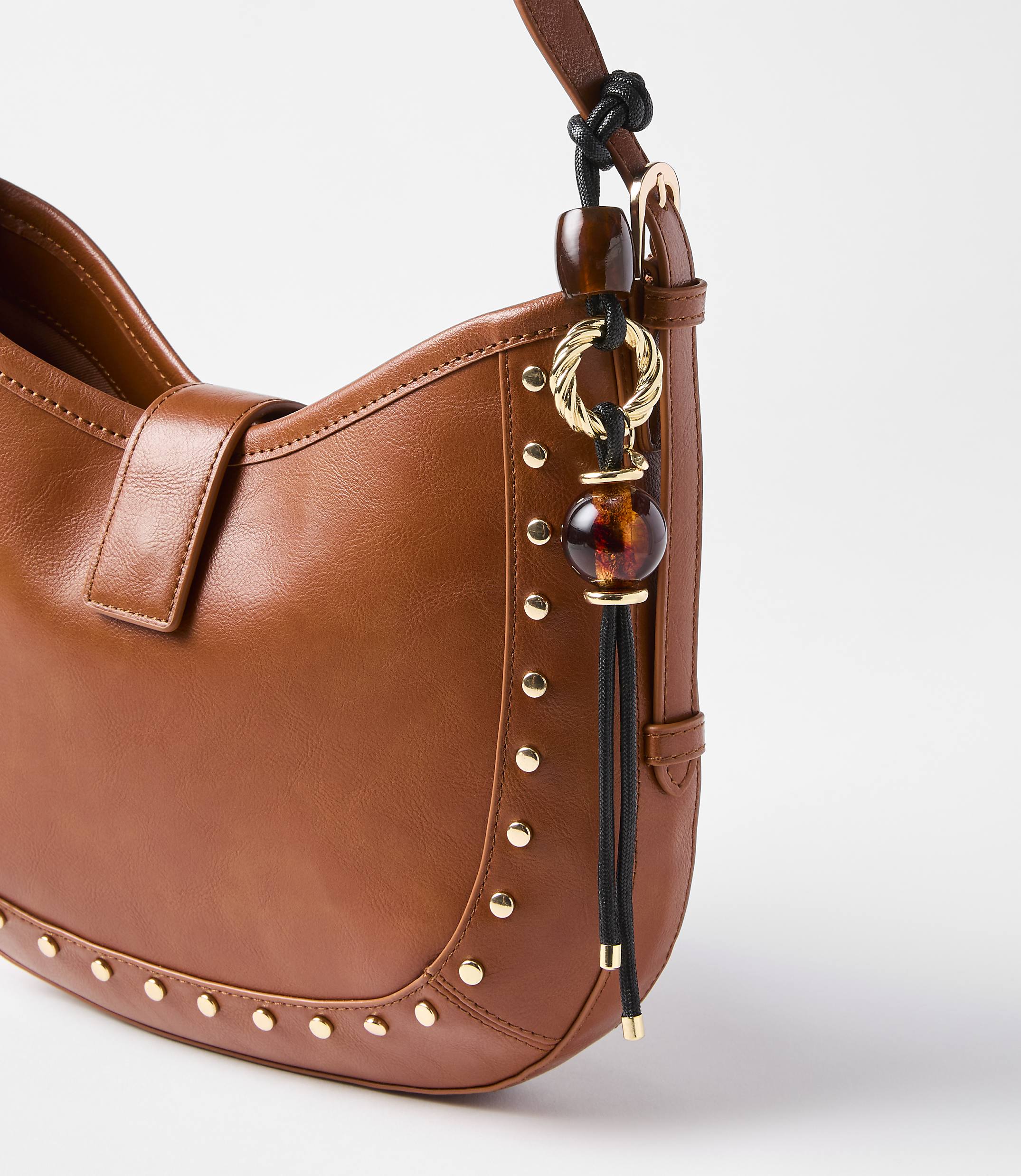 A close-up of a brown leather handbag with gold stud detailing and a decorative charm hangs against a plain white background.