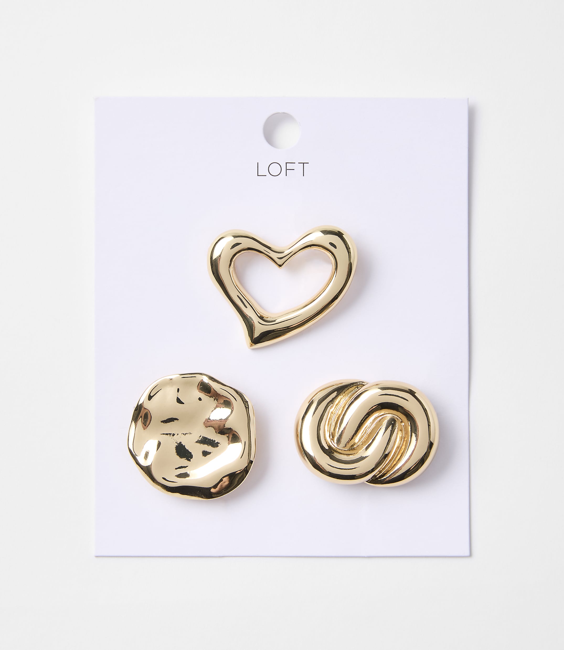 Three gold-toned charms, a heart, a textured circle, and a knot, are displayed on a white card with the "LOFT" logo against a plain white background.