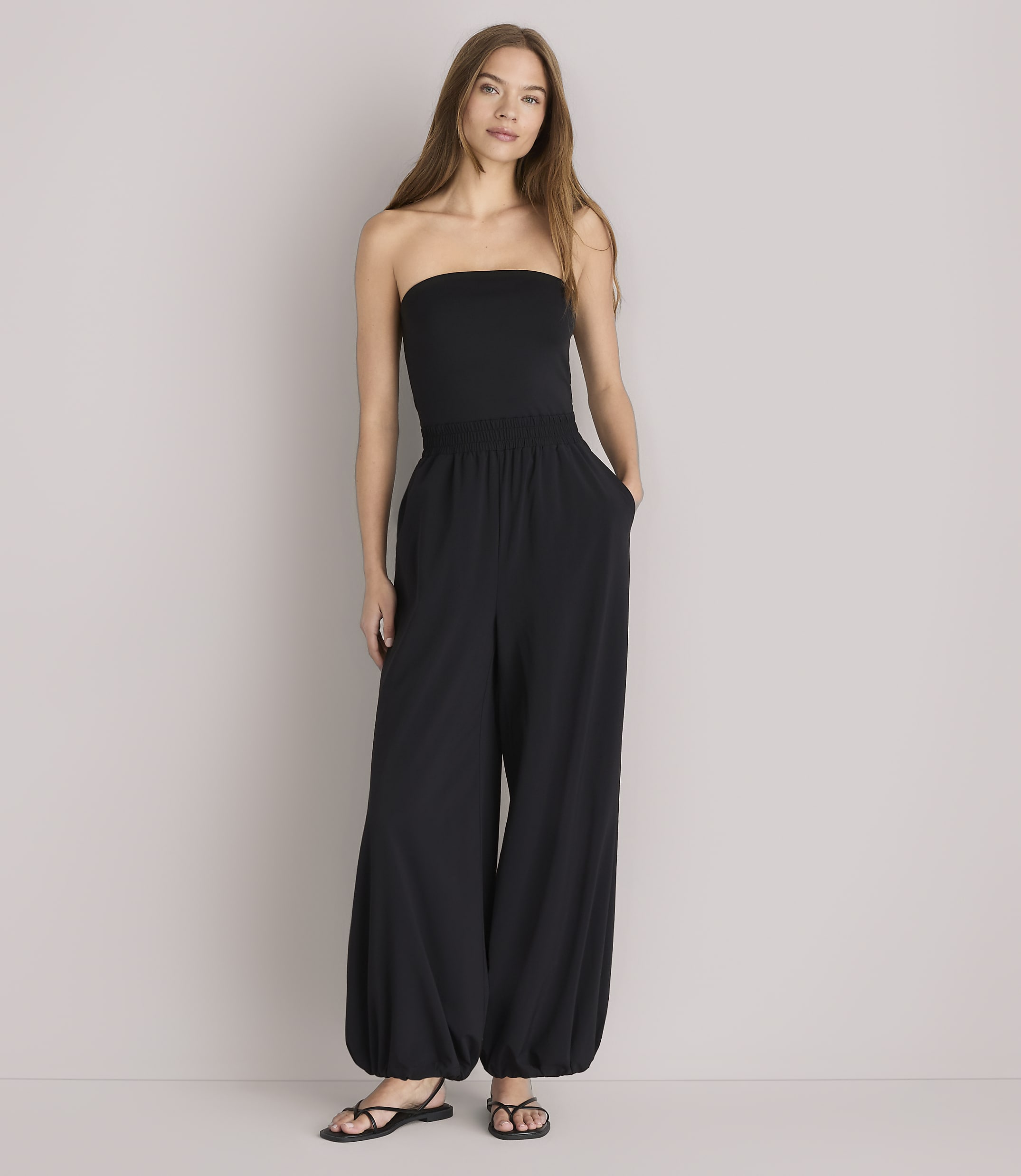 A woman in a black strapless jumpsuit and white open shirt stands against a plain white background.