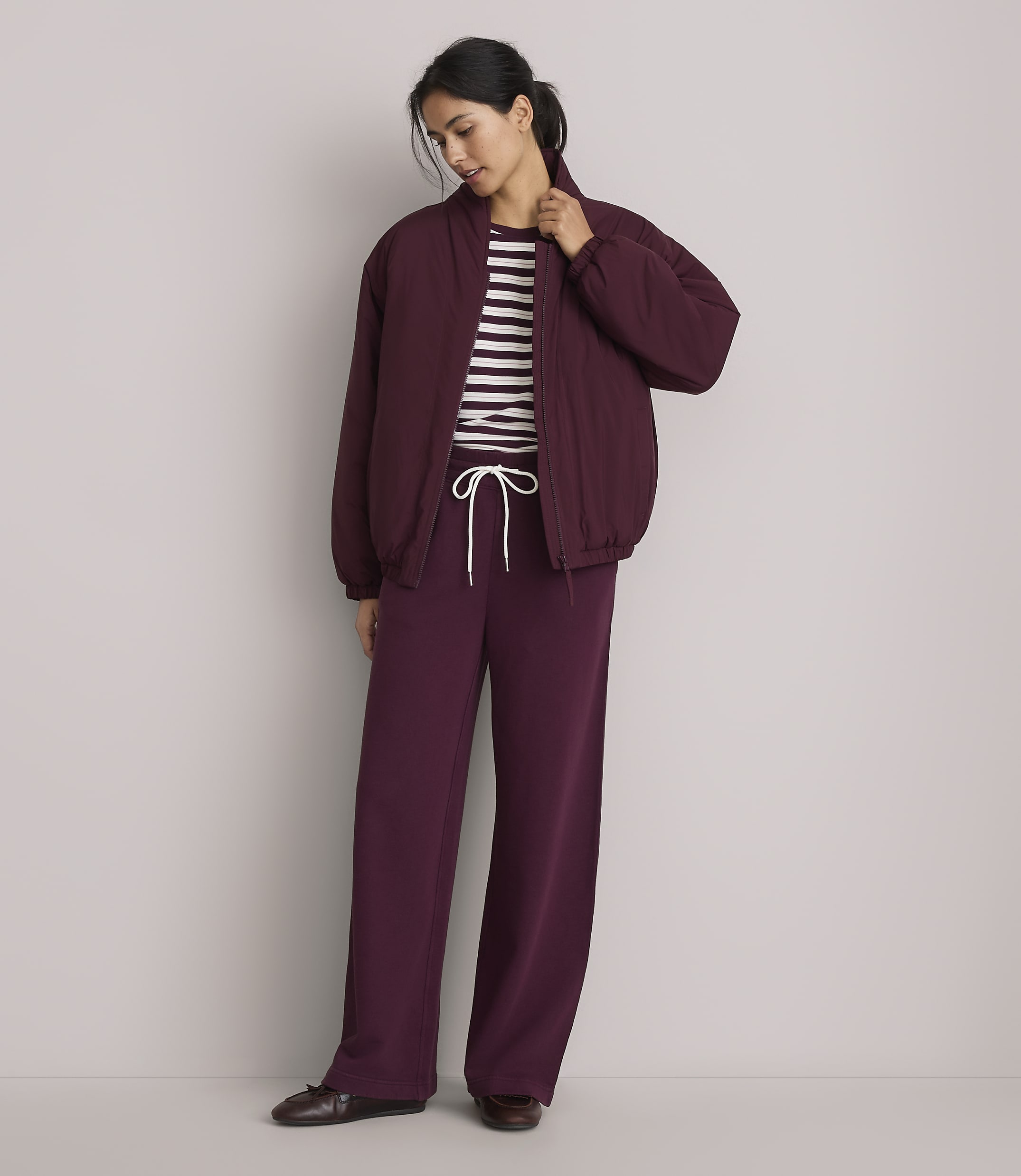 A woman in a burgundy bomber jacket and matching wide-leg pants stands against a plain gray background.