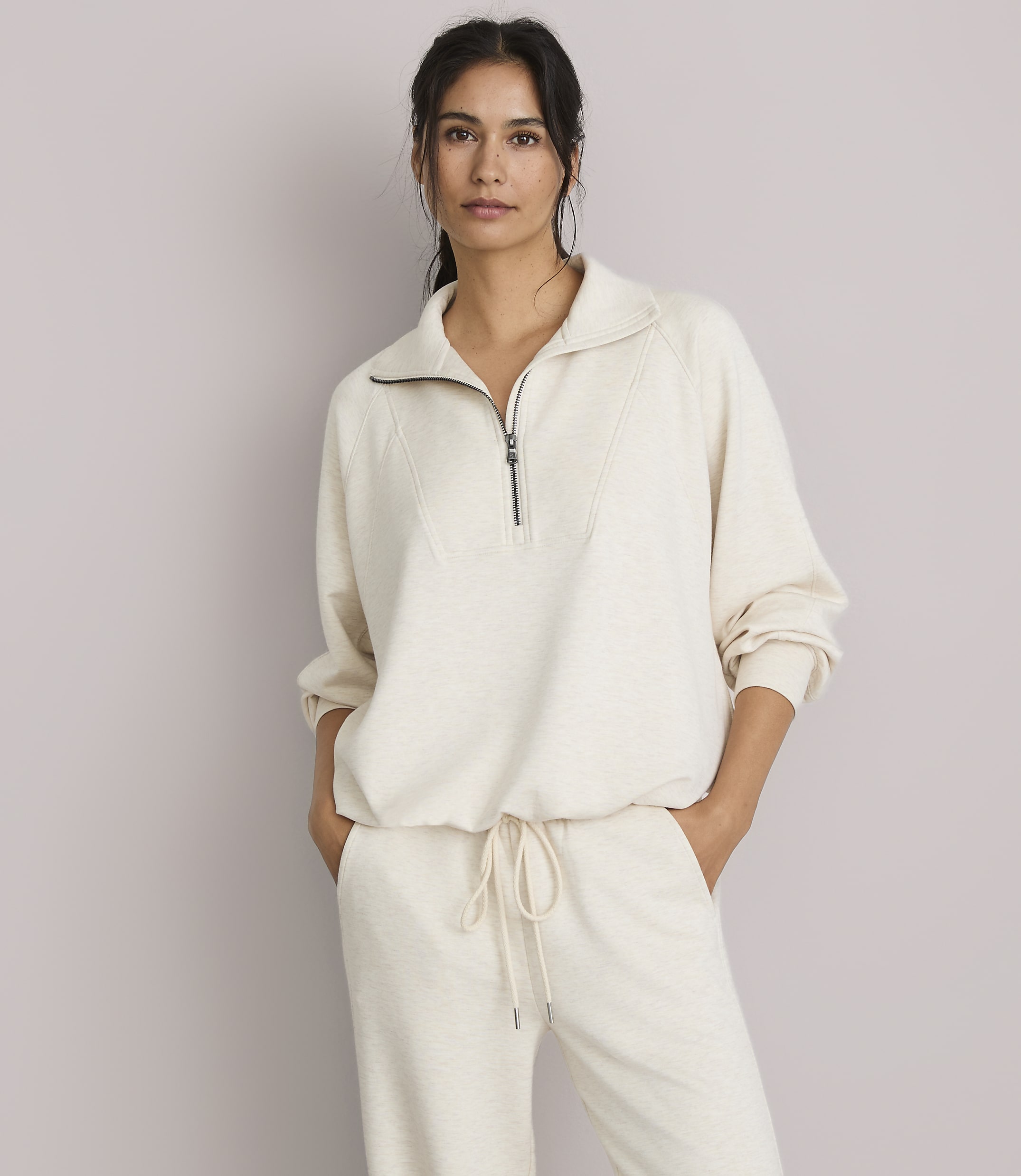 A woman in a cream-colored quarter-zip sweatshirt and matching pants stands against a plain, light gray background.