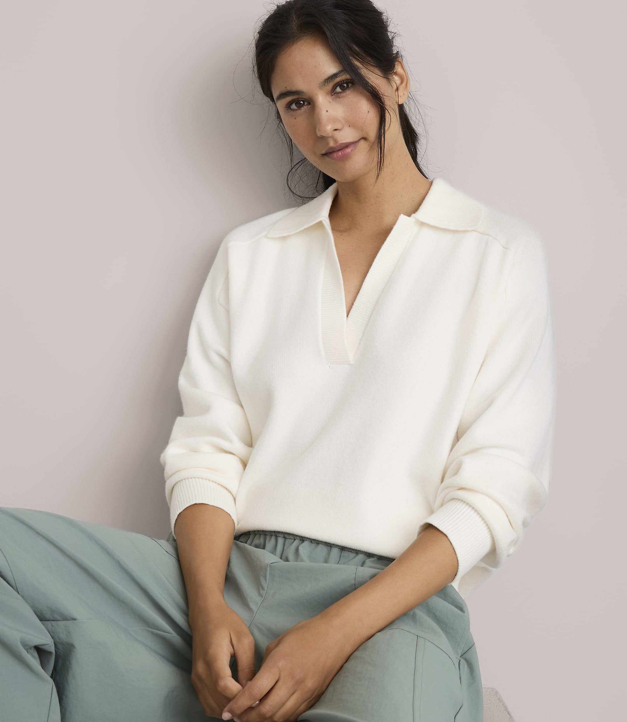 A woman in a cream-colored collared sweater and light green pants sits against a plain, light-colored background.