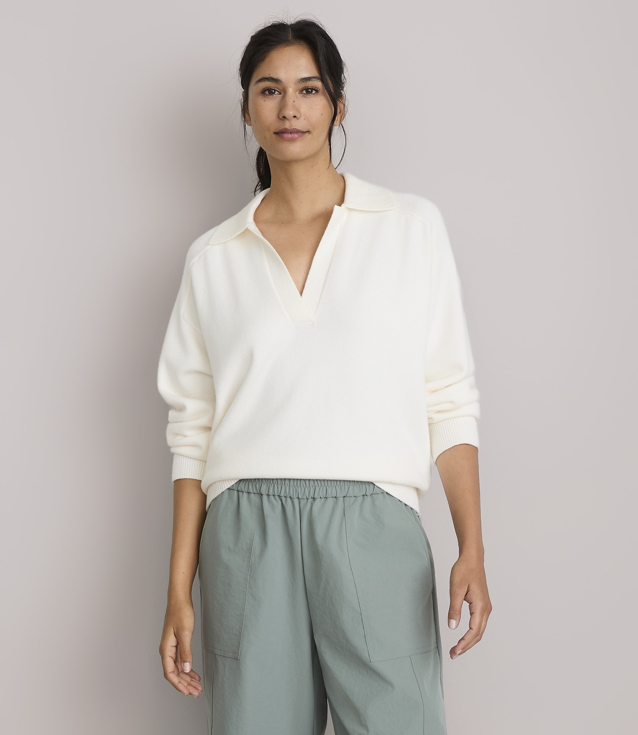 A woman in a cream-colored collared sweater and green pants stands against a plain, light gray background.