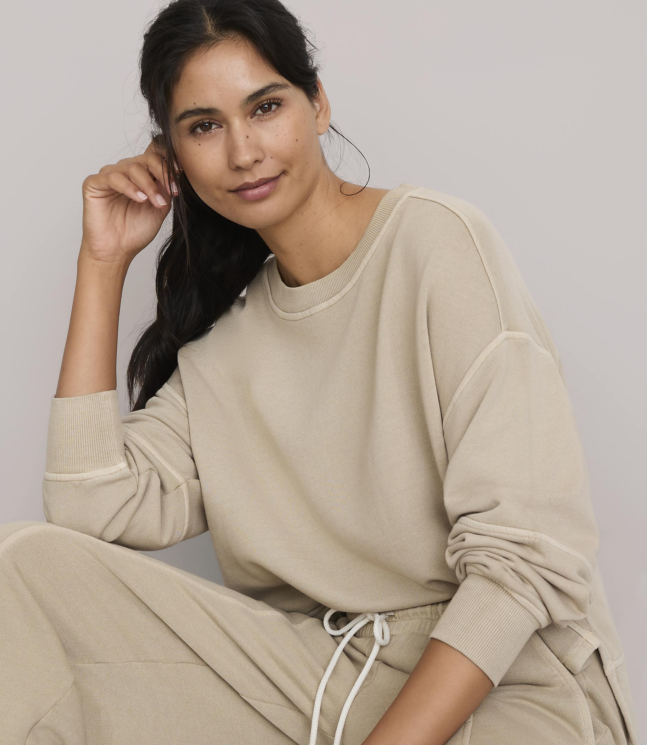 A woman in a beige sweatshirt and pants poses against a plain, light background.