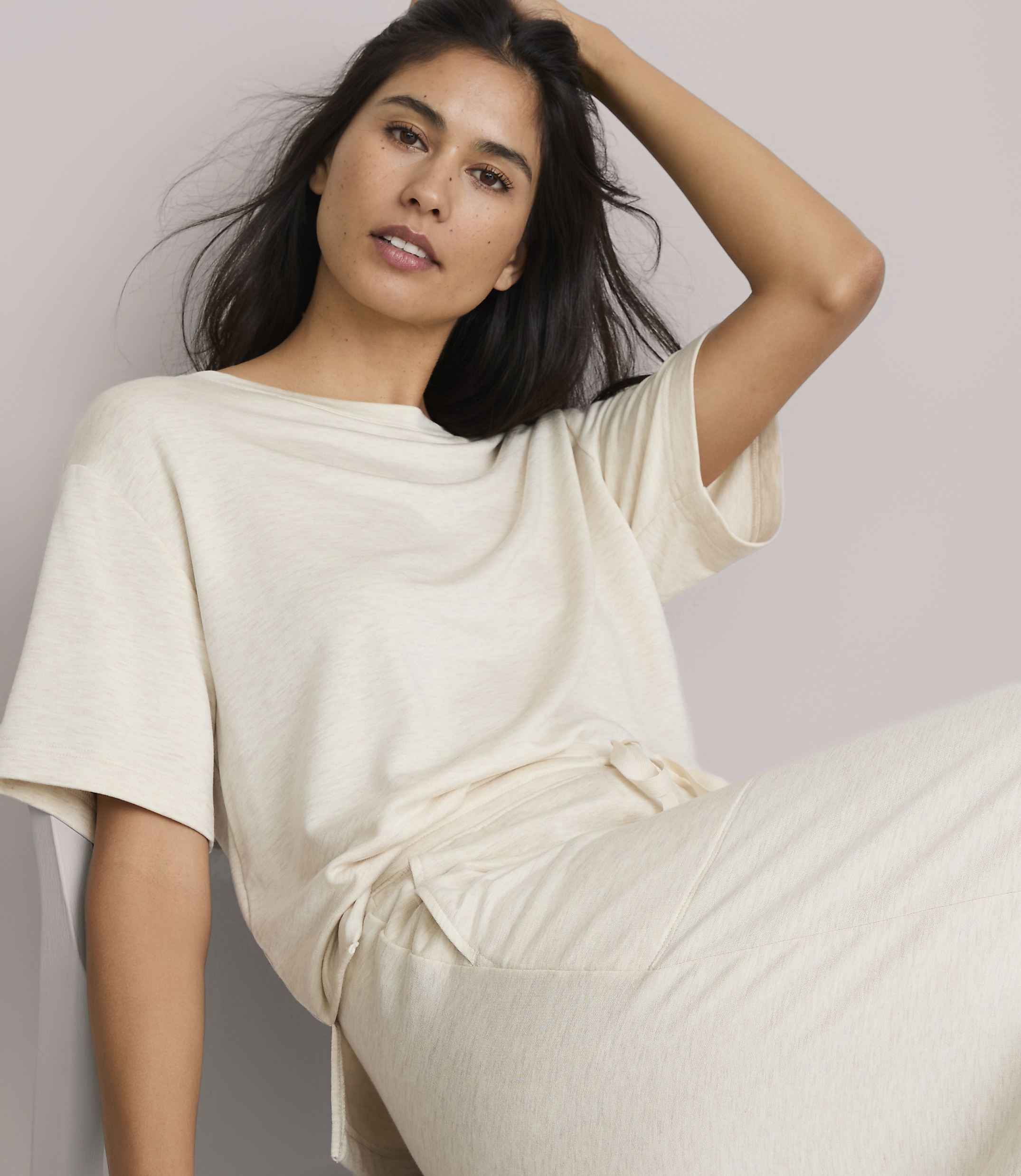 A woman in a cream-colored t-shirt and pants poses against a plain, light-colored background.