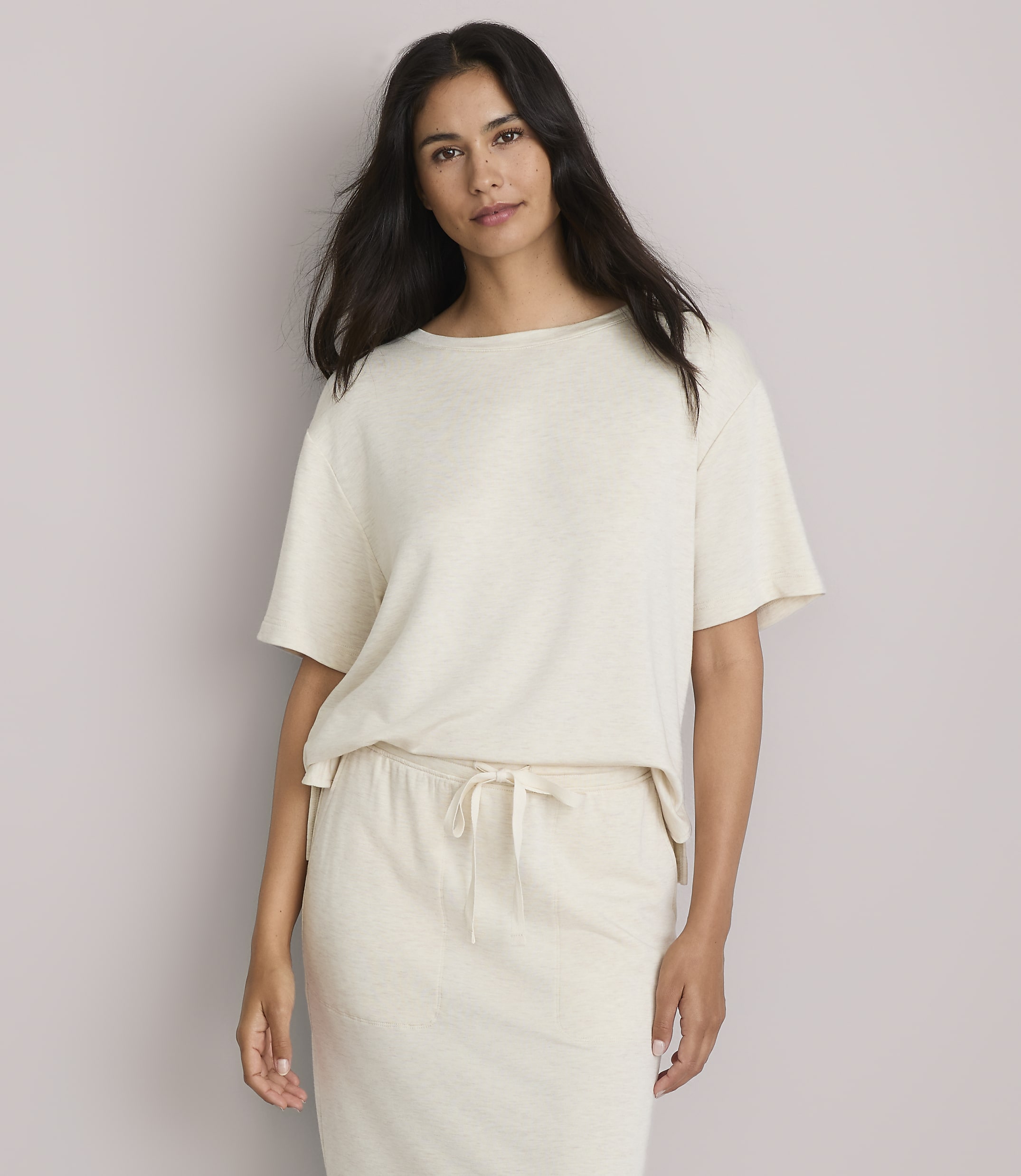 A woman in a cream-colored t-shirt and matching pants stands against a plain, light gray background.