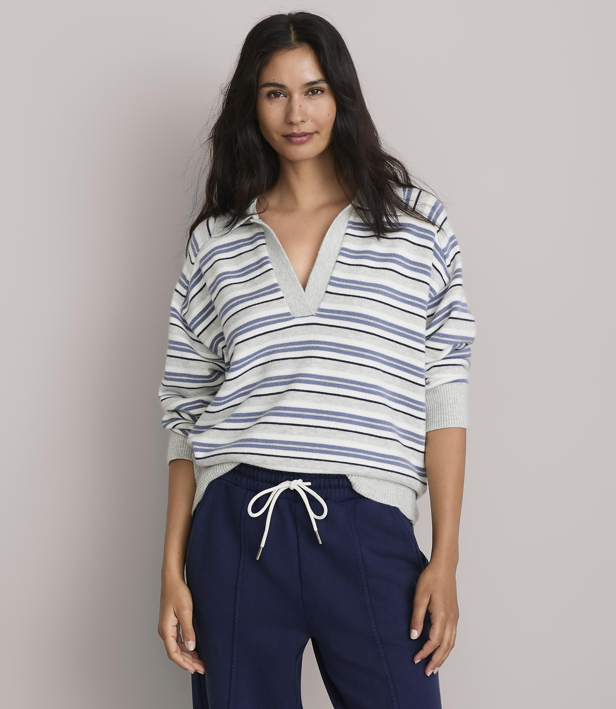 A woman in a striped collared shirt and navy sweatpants stands against a plain gray background.