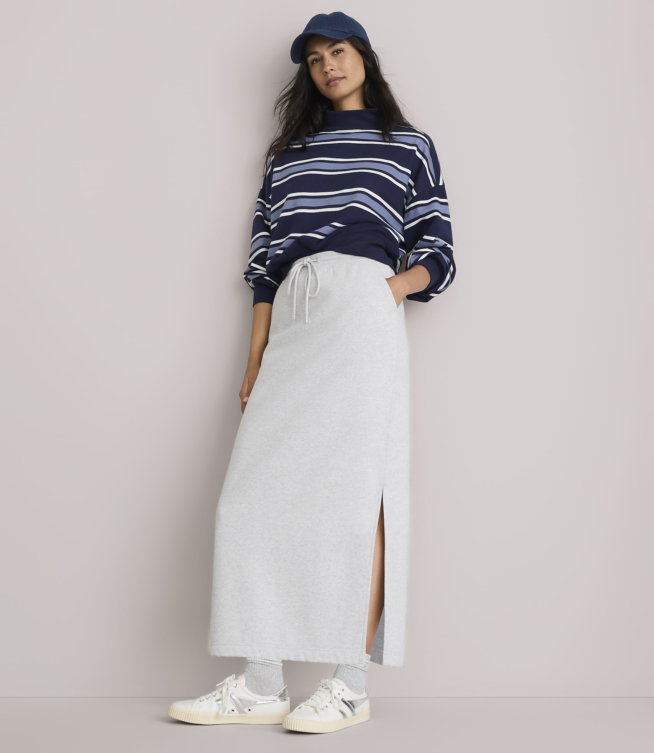 A woman in a striped sweatshirt and a light gray maxi skirt with a side slit stands against a plain, light gray background.