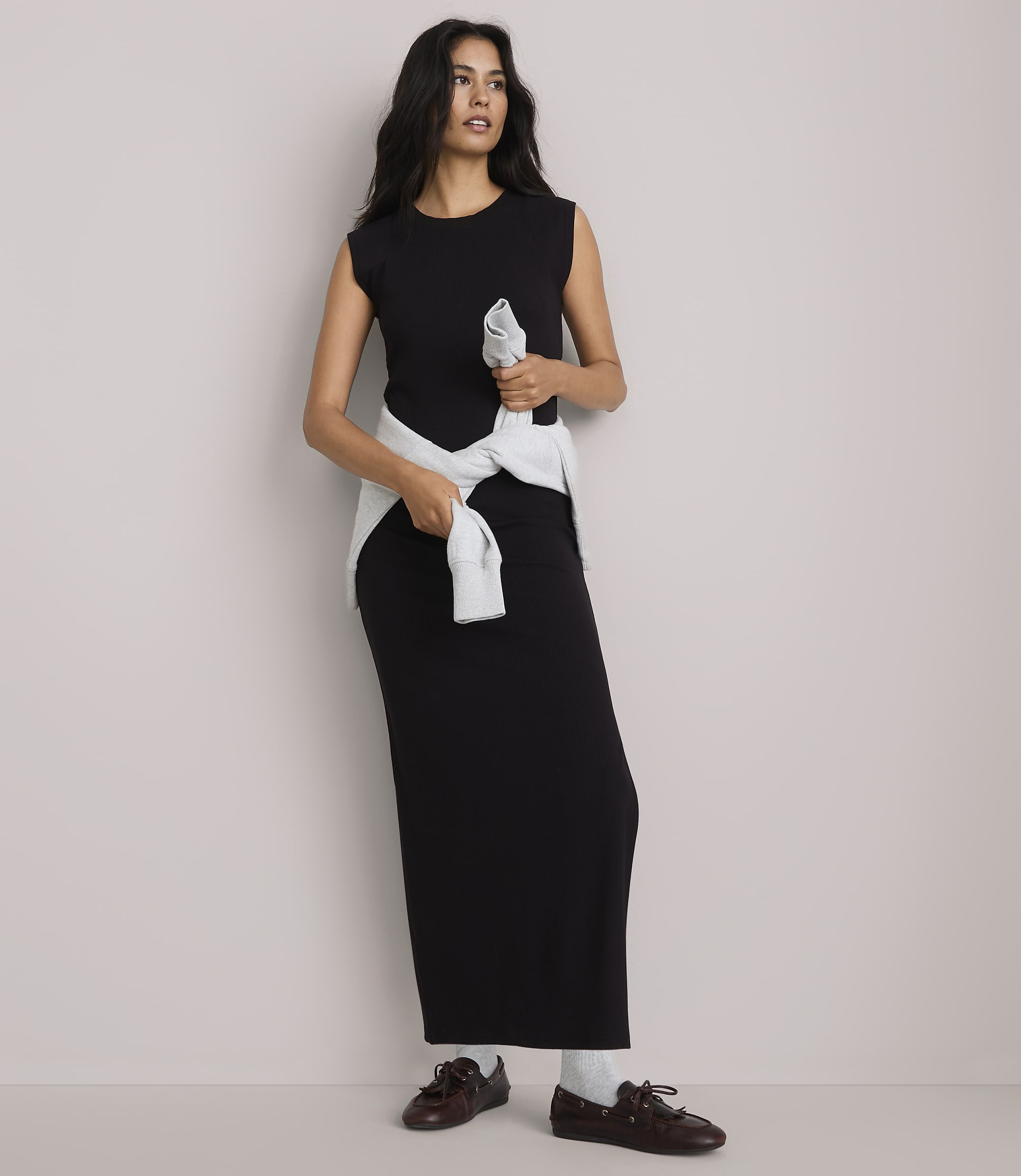 A woman in a black maxi dress and light blue sweater tied around her waist stands against a plain, light gray background.