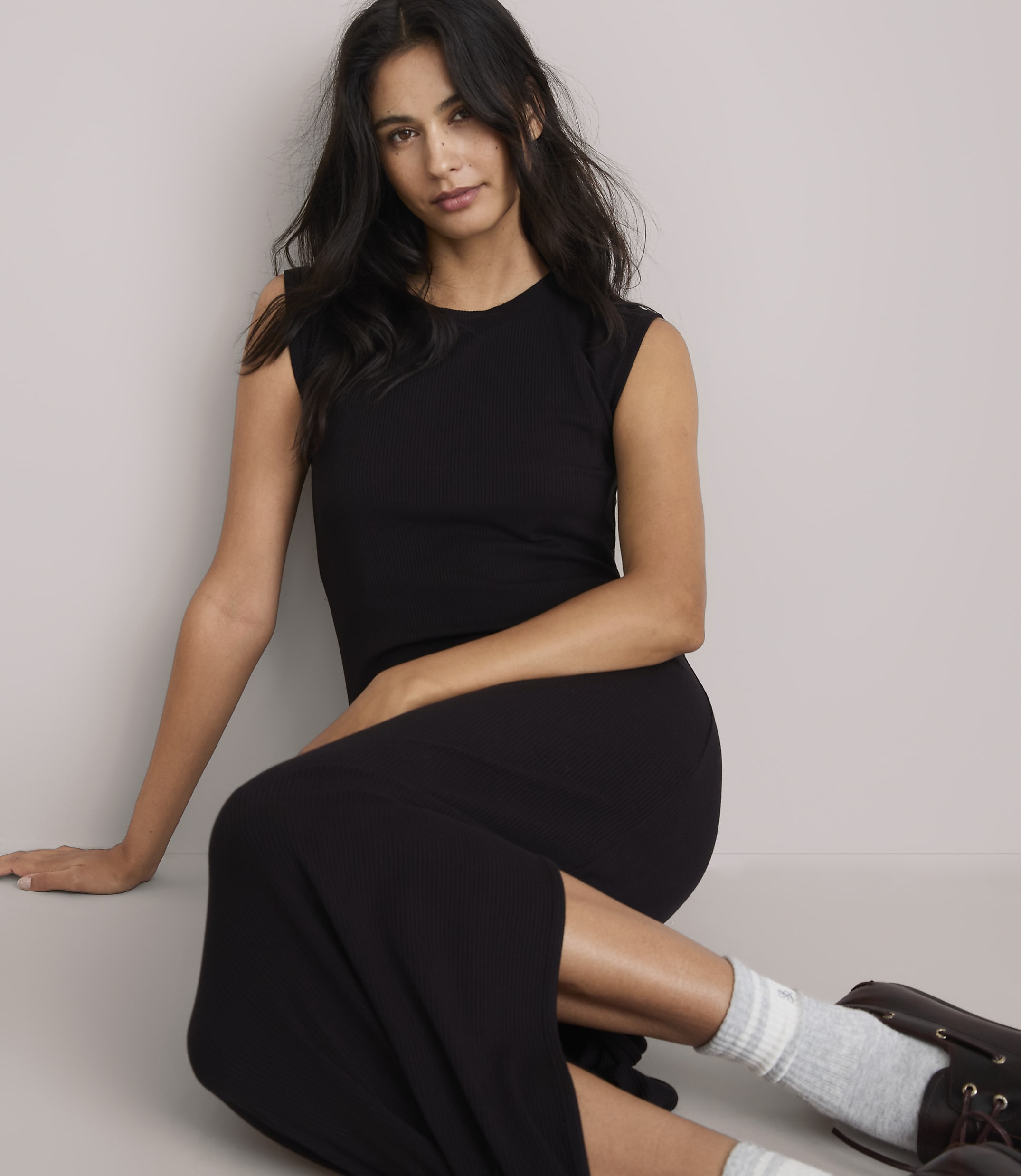 A woman in a black ribbed dress and grey socks poses on a light grey floor with a plain grey background.