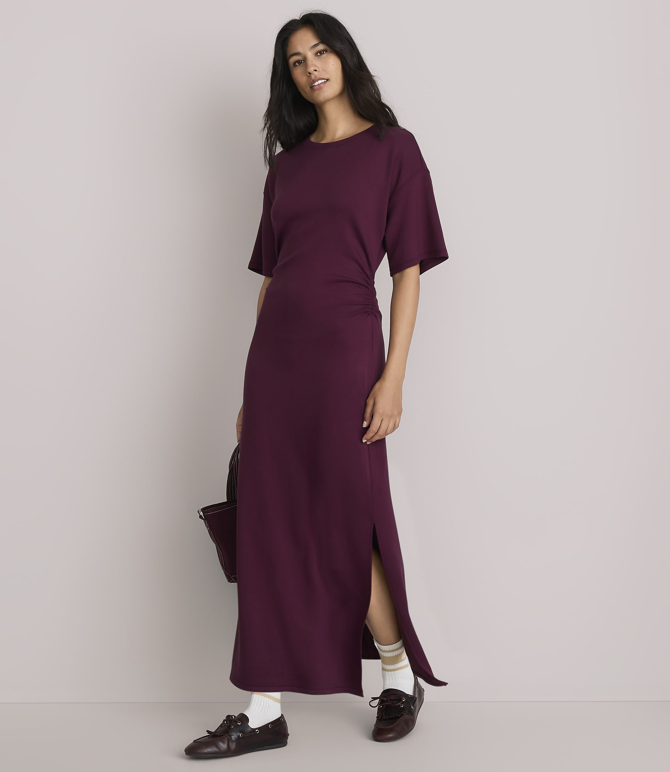 A woman in a plum-colored maxi dress with a side slit stands against a plain, light gray background, holding a dark bag.