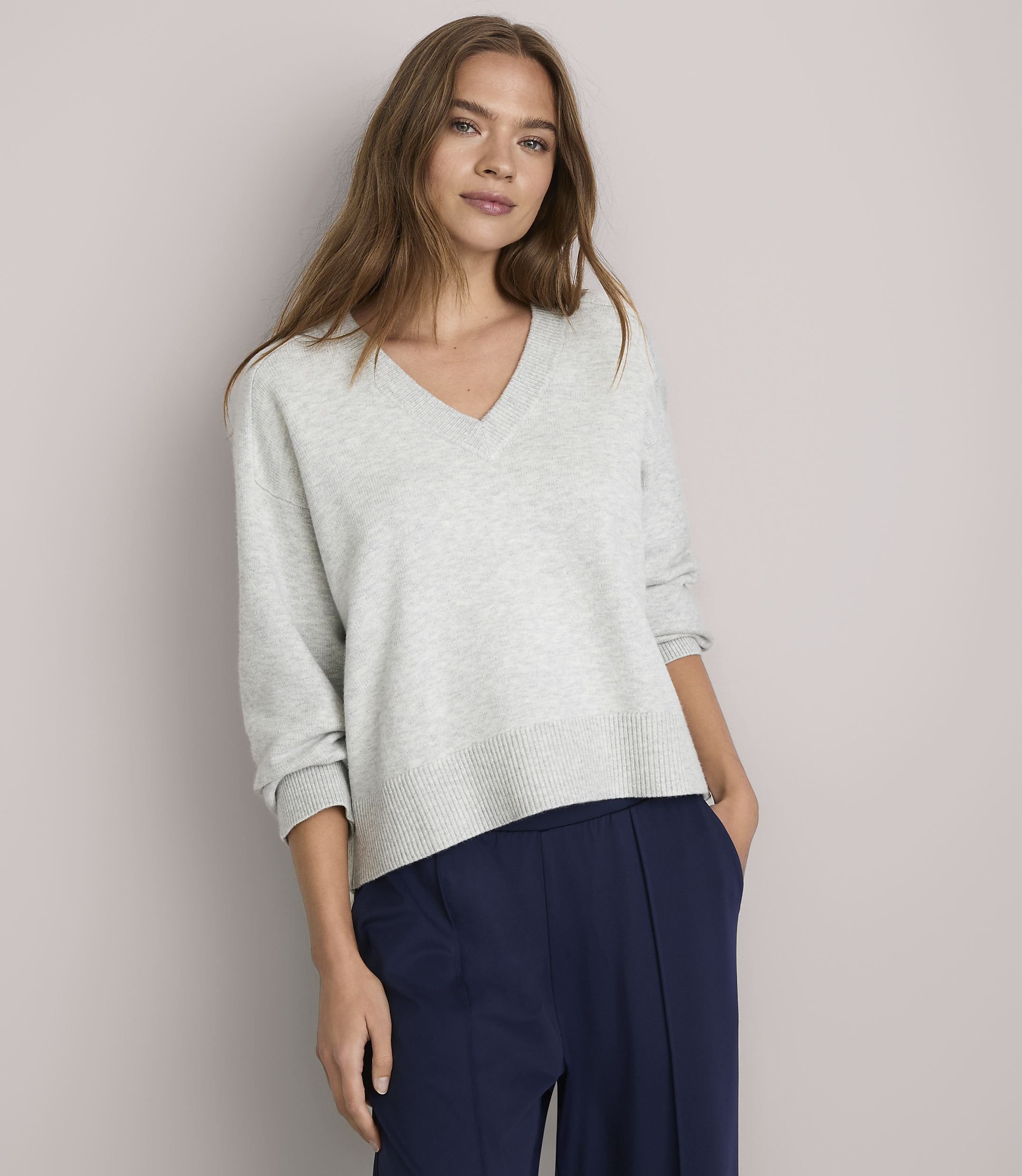 A woman in a light gray v-neck sweater and navy blue pants stands against a plain white background.