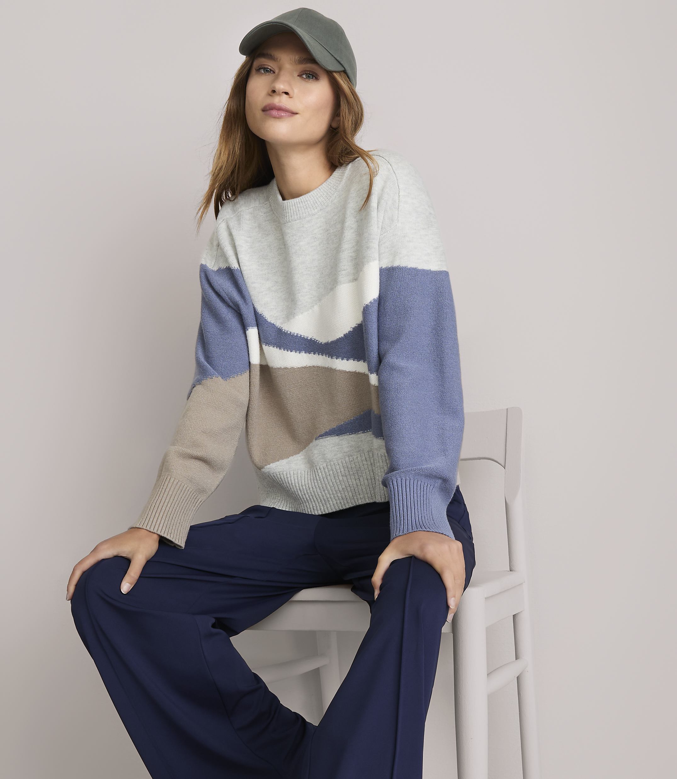A woman in a patterned sweater and navy pants sits on a white chair against a plain white background.