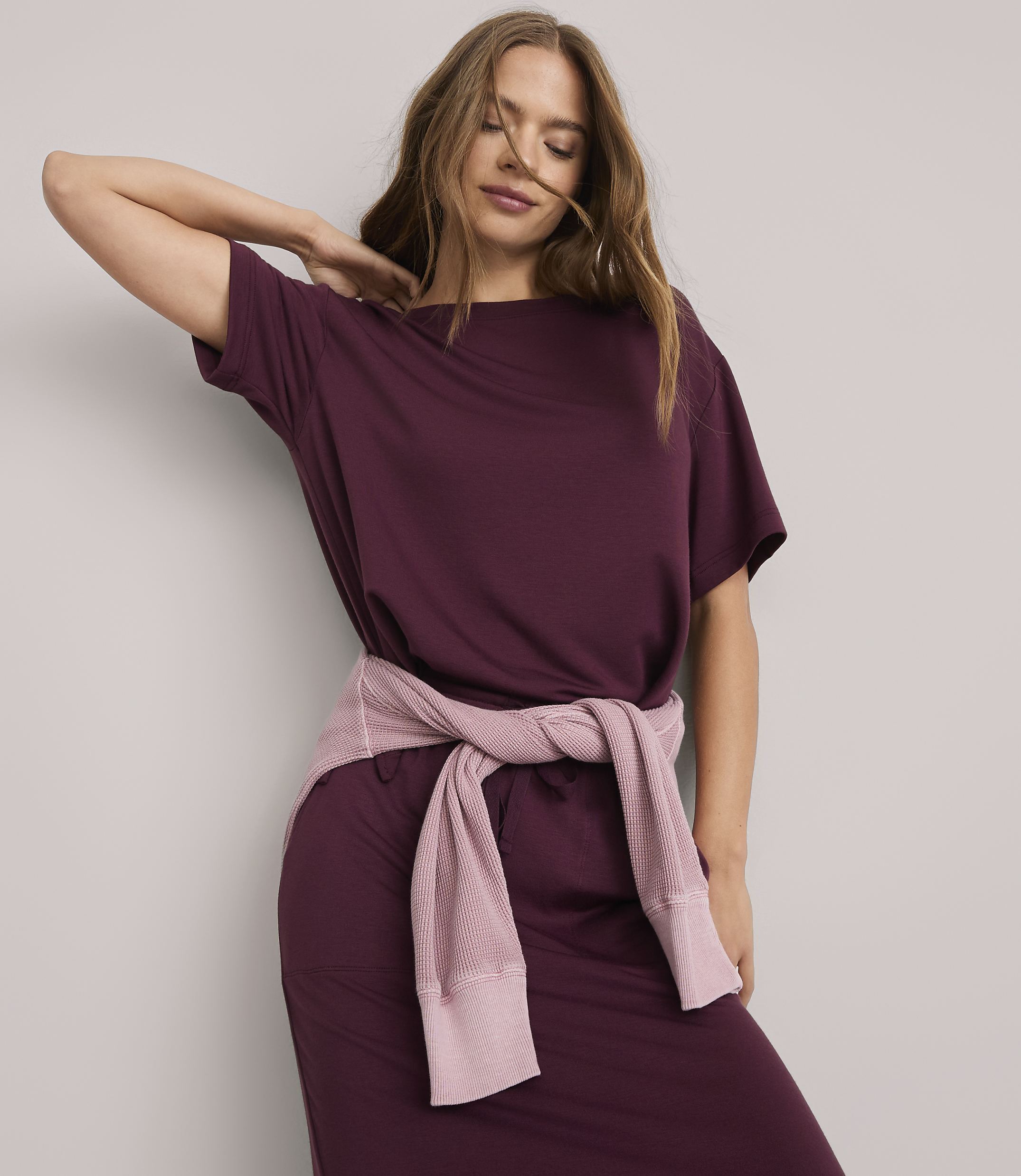 A woman in a burgundy dress with a pink sweater tied around her waist poses against a plain white background.