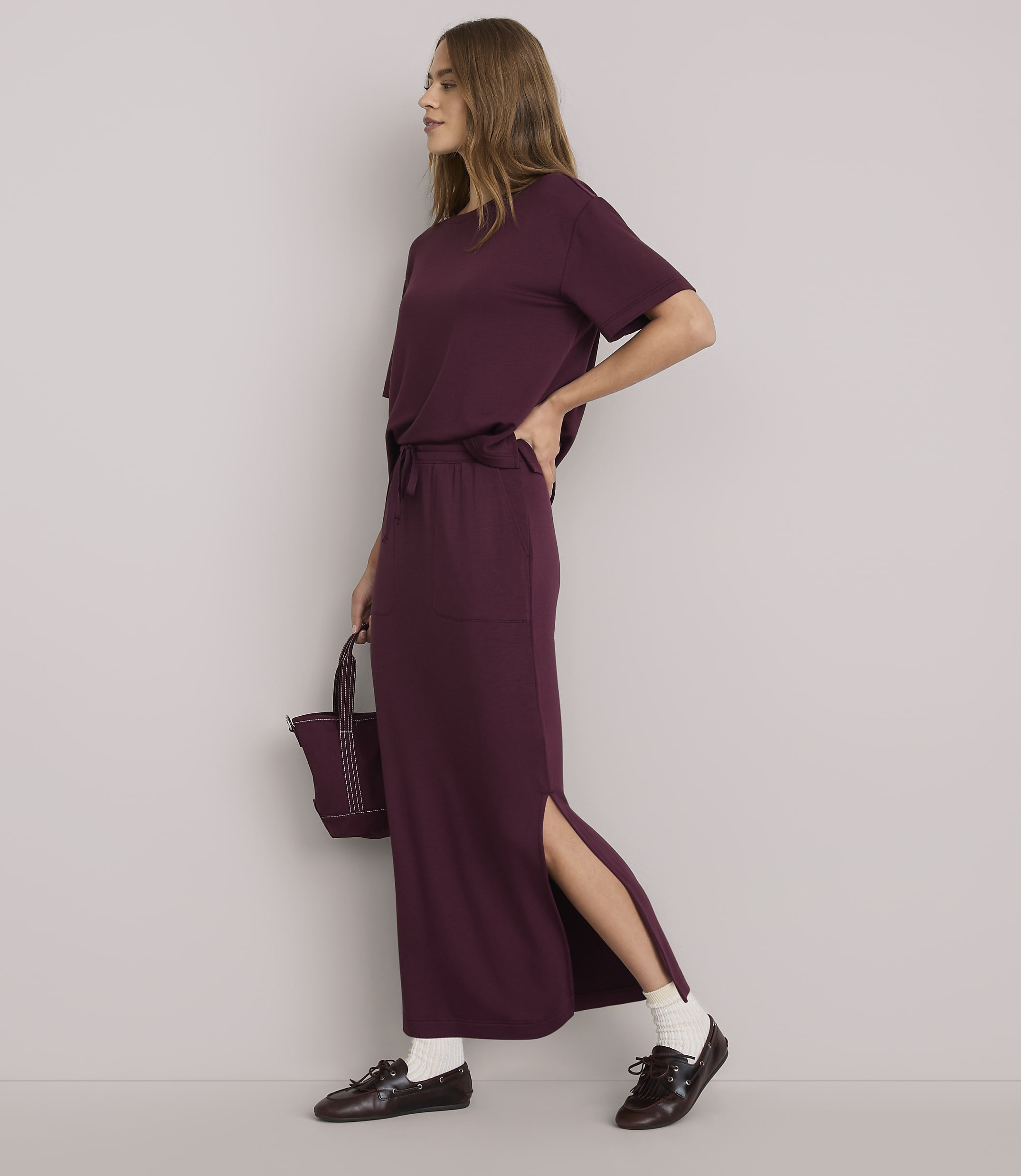 A woman in a burgundy t-shirt and matching maxi skirt with a side slit stands against a plain white background, holding a small burgundy bag.
