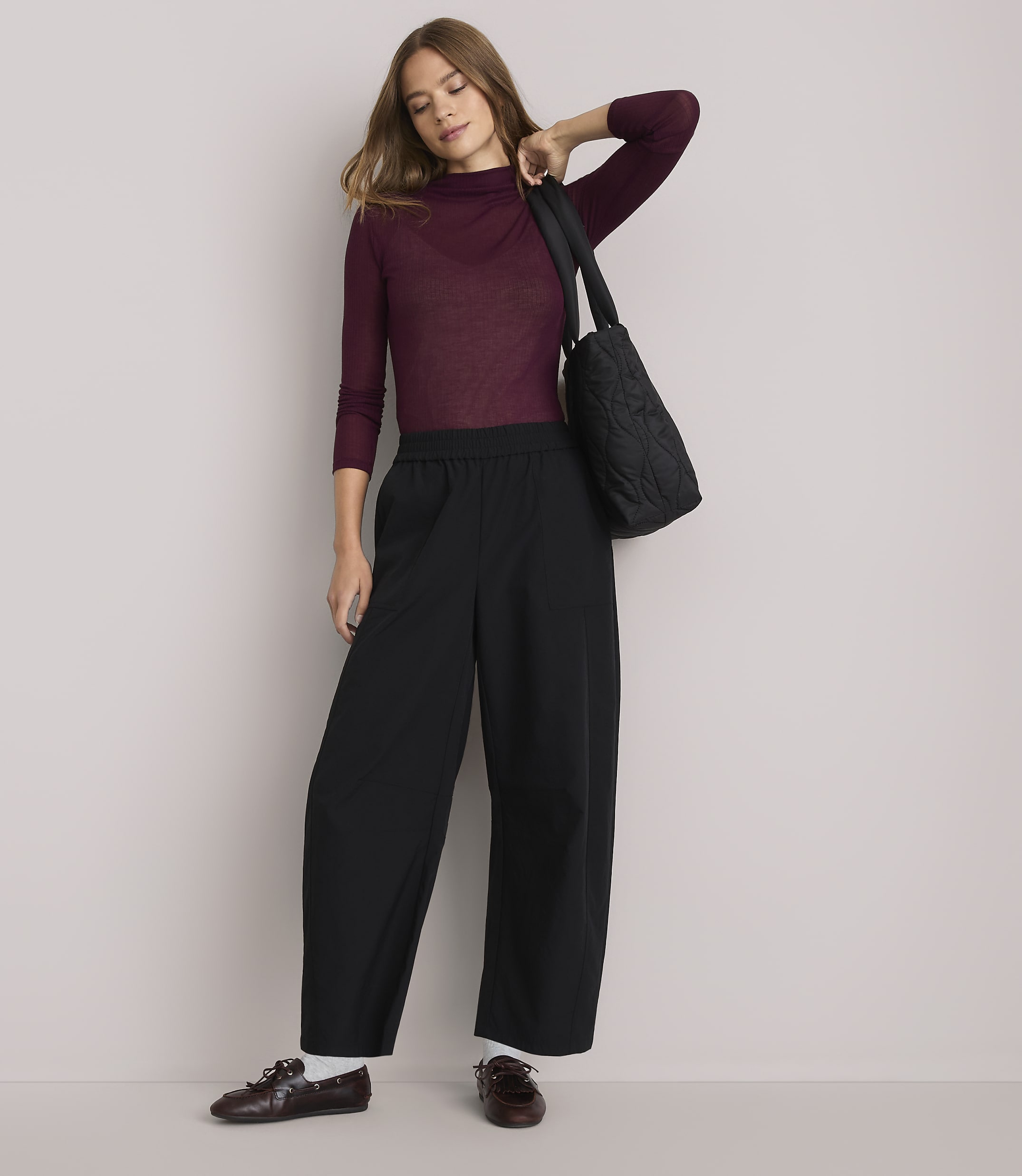A woman in a burgundy turtleneck and black wide-leg pants stands against a plain white background, carrying a quilted black tote bag.