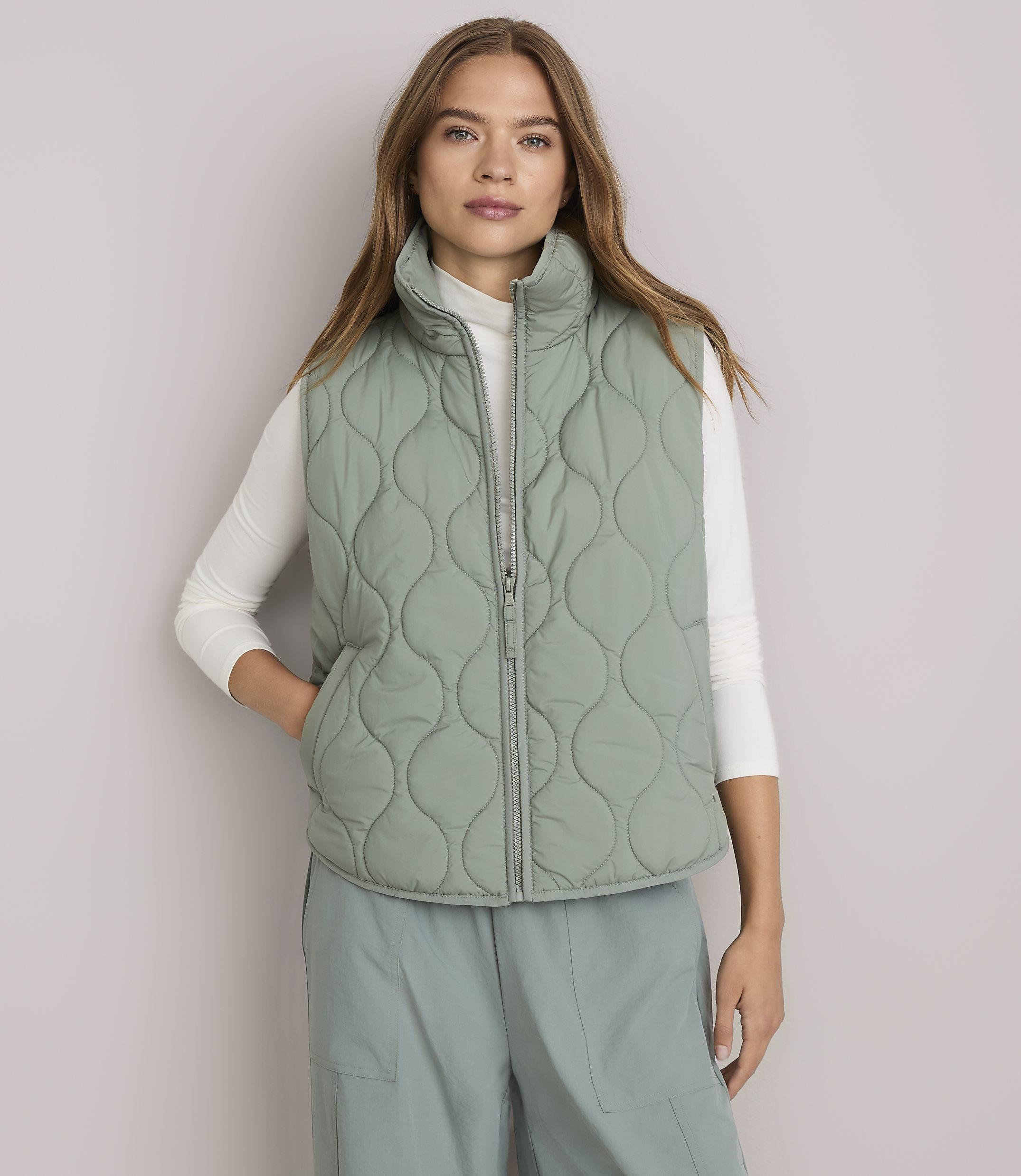 A woman in a light green quilted vest and white turtleneck stands against a plain white background.