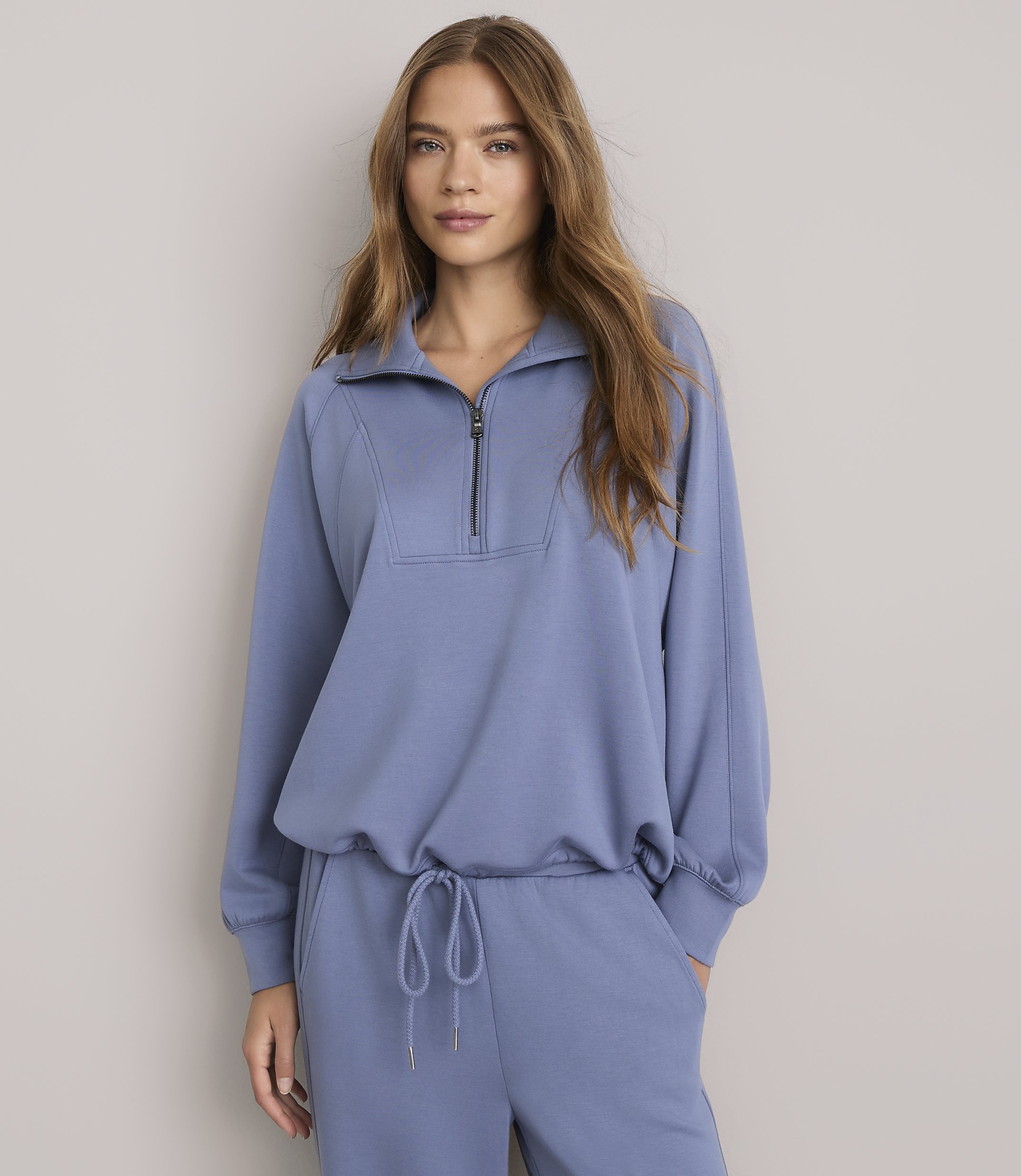 A woman in a blue quarter-zip sweatshirt and matching sweatpants stands against a plain gray background.