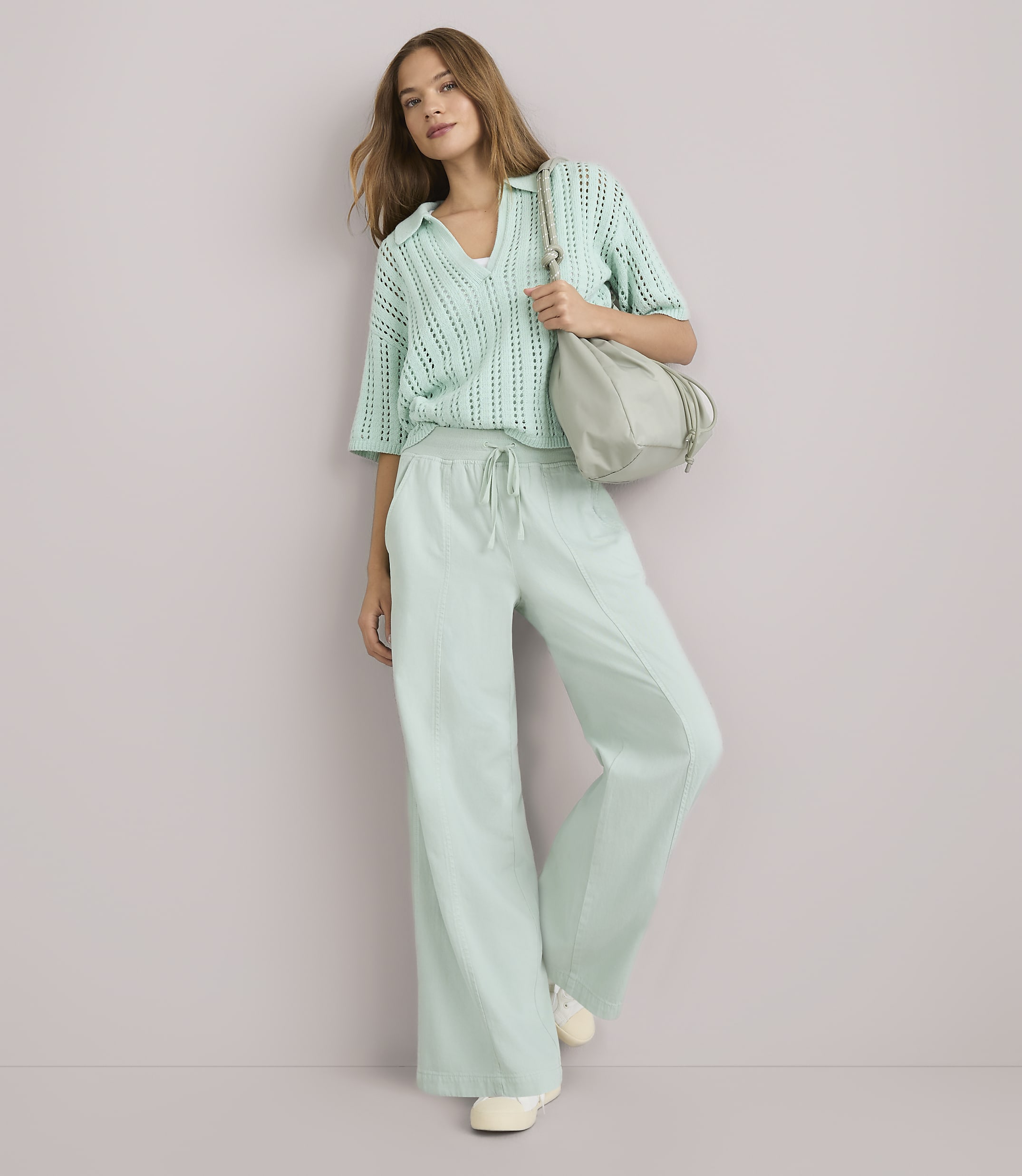 A woman in a mint green knit top and wide-leg pants stands against a plain white background, carrying a light gray bag.