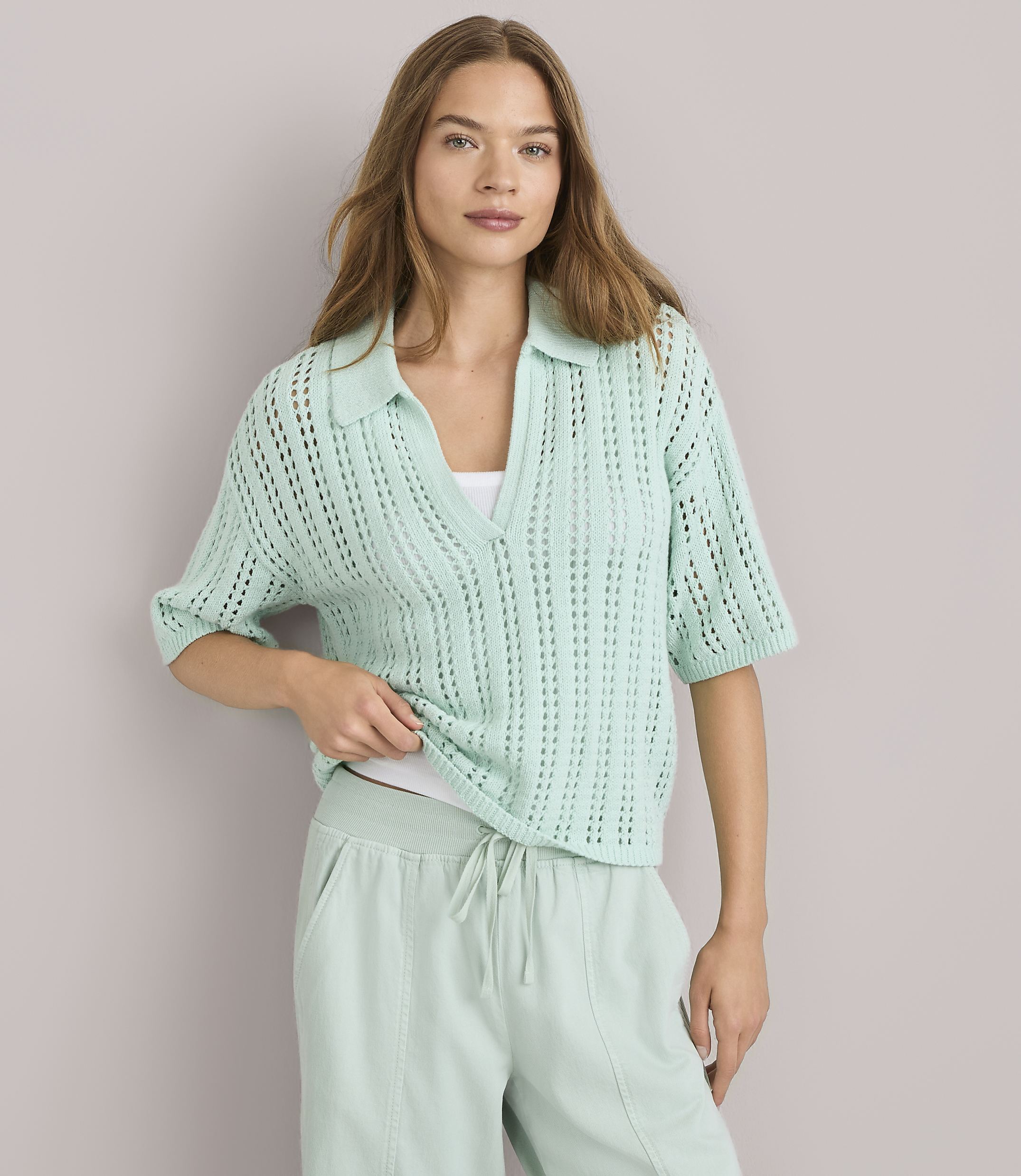 A woman in a light green collared knit shirt and matching sweatpants stands against a plain gray background.