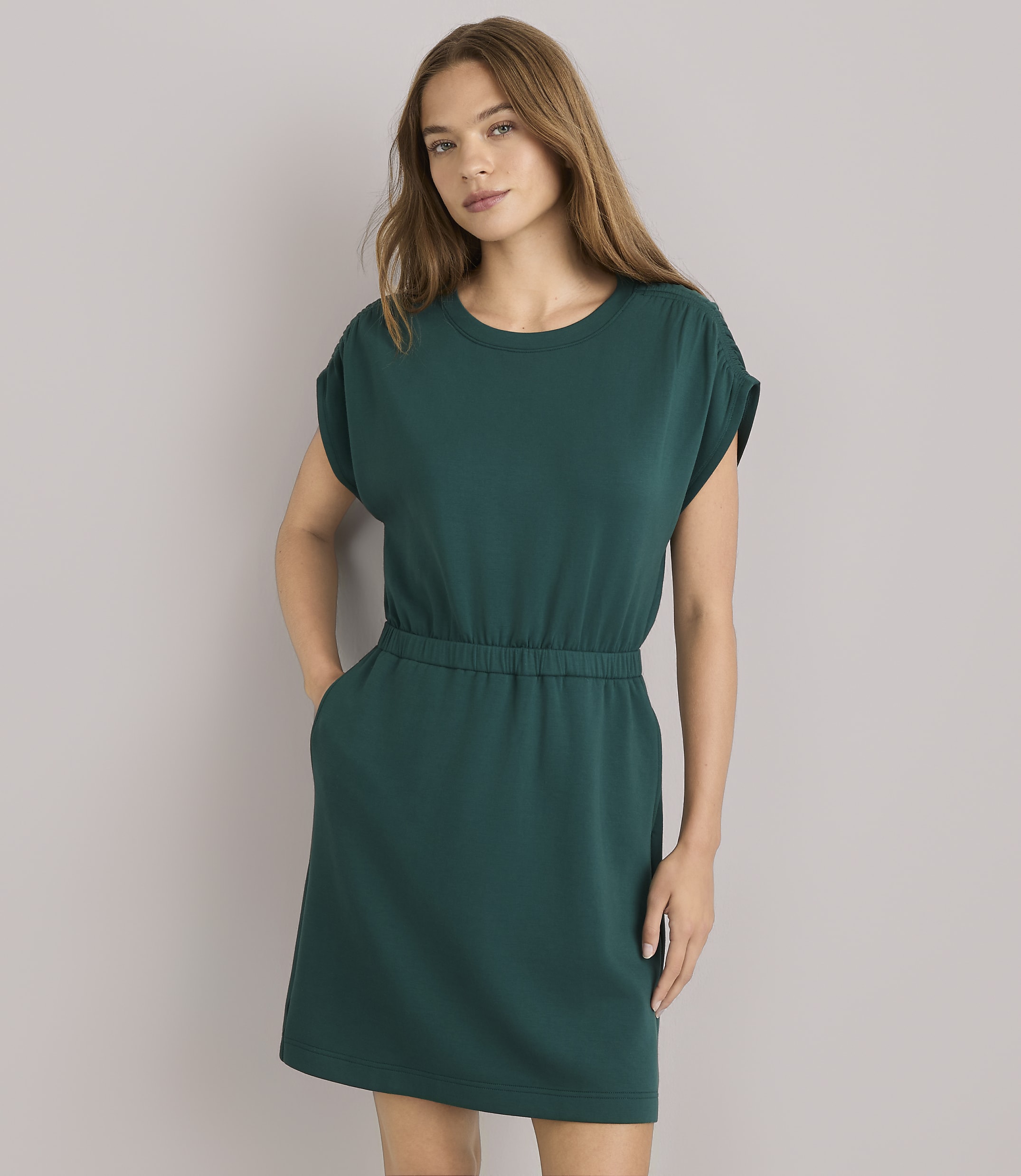 A woman in a green dress and sunglasses leans against a plain gray wall, with a light blue sweater tied around her waist.