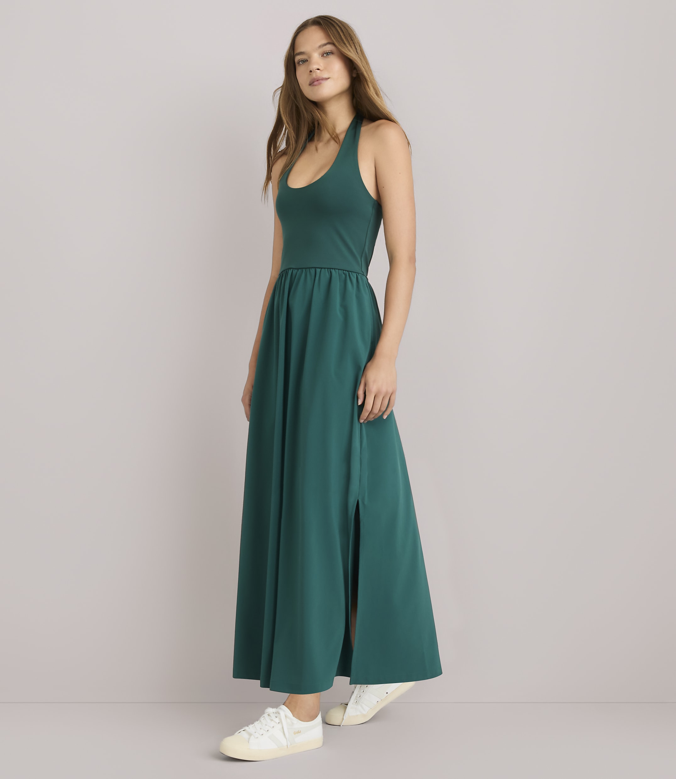 A woman in a teal maxi dress with a high slit and white sneakers stands against a plain gray background.