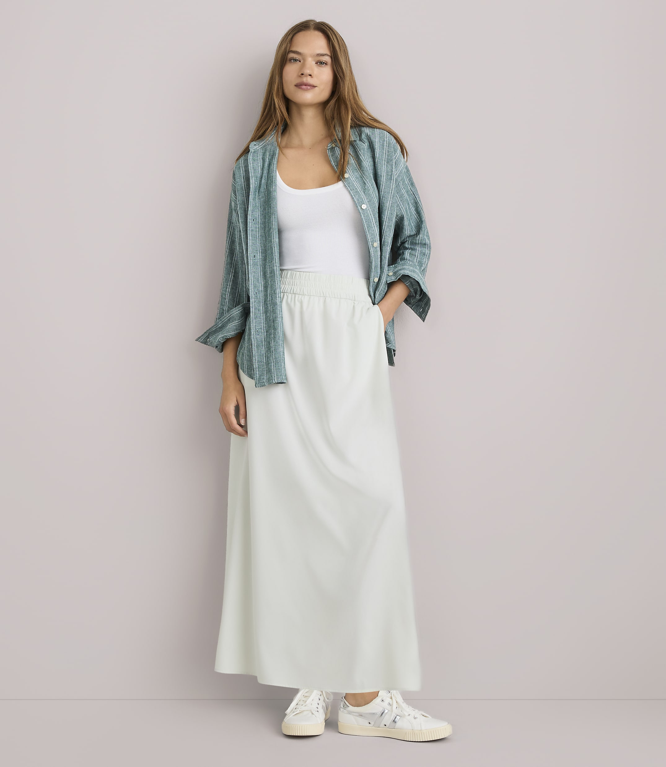 A woman in a white tank top, a green patterned shirt, and a long white skirt stands against a plain white background.