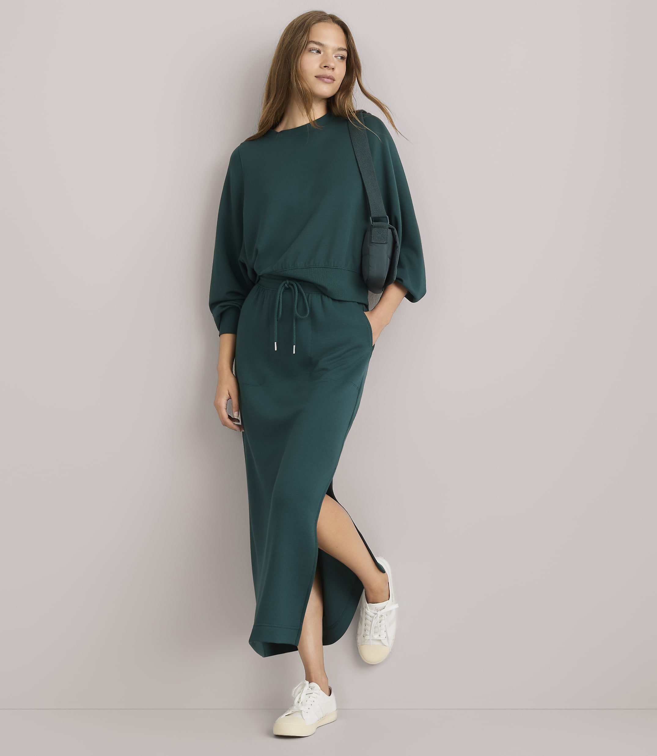 A woman in a teal sweatshirt and matching skirt with a side slit stands against a plain white background, wearing white sneakers and a small crossbody bag.