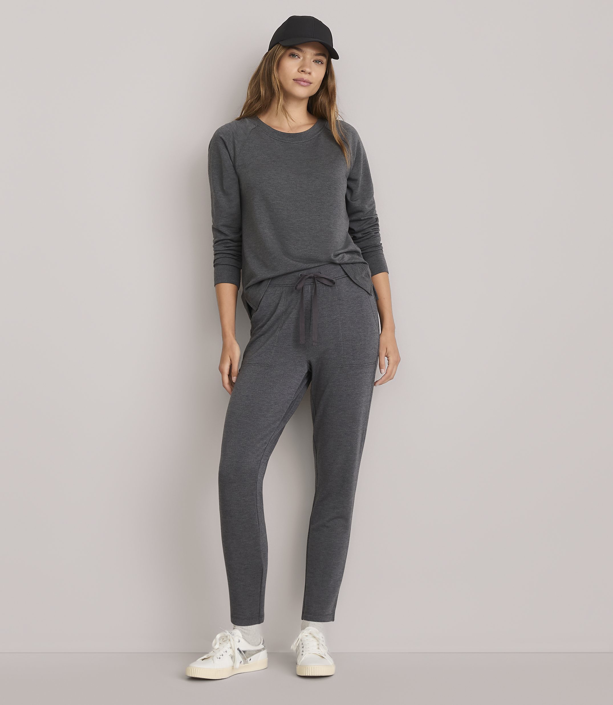 A woman in a dark gray loungewear set and a black baseball cap stands against a plain white background.