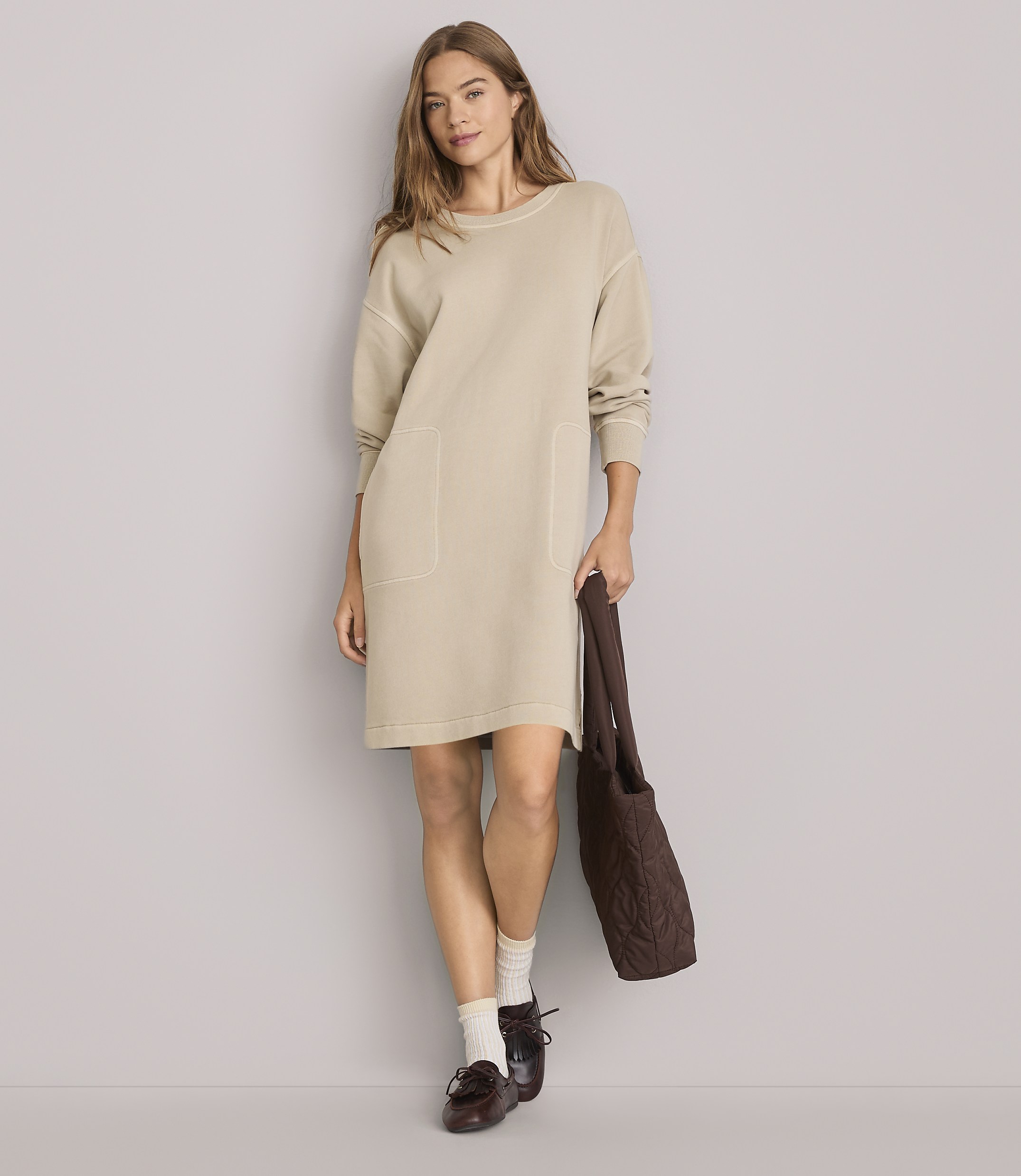 A woman in a beige dress and loafers stands against a plain gray background, holding a brown tote bag.