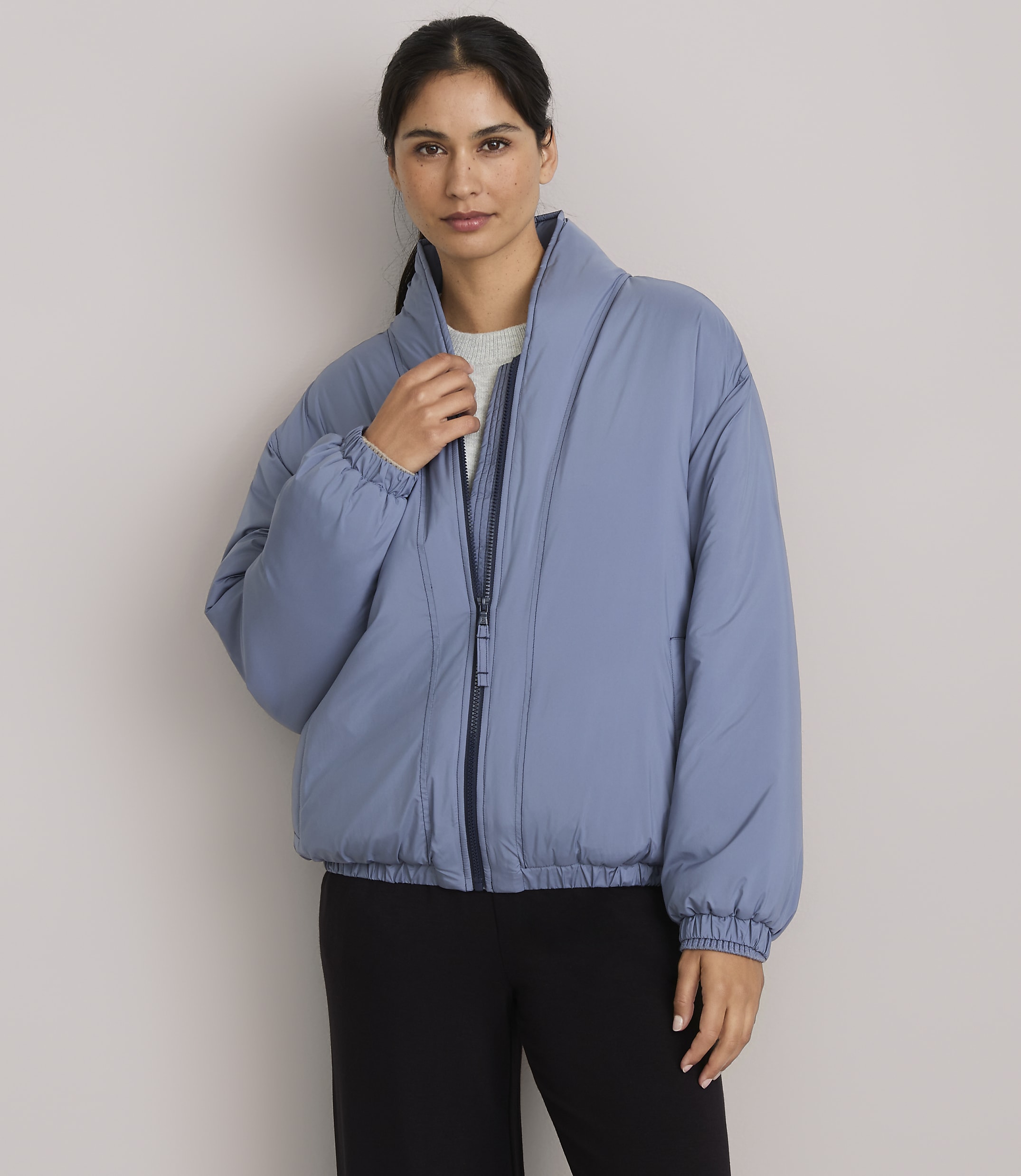A woman in a blue puffer jacket stands against a plain gray background.