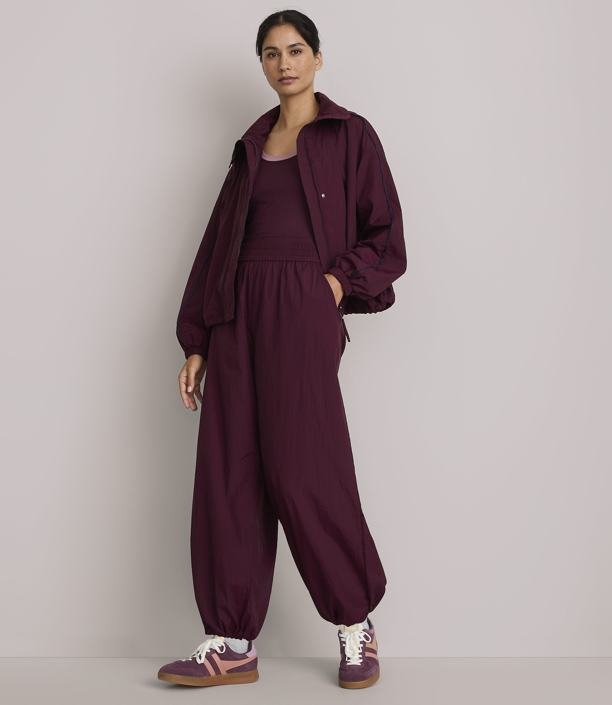 A woman in a burgundy tracksuit stands against a plain gray background.