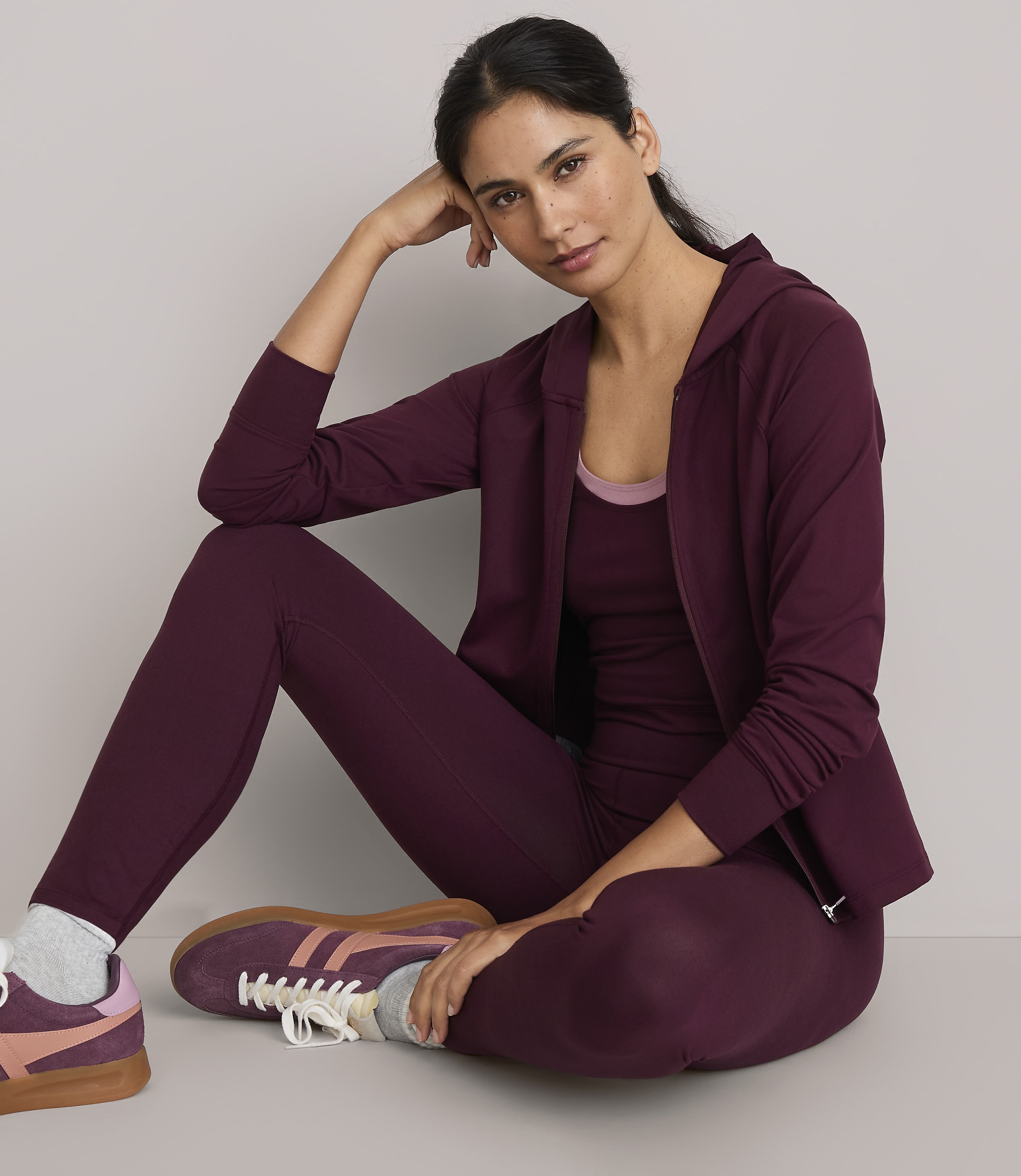 A woman in burgundy activewear sits on a plain gray floor with a matching hoodie and leggings, her hand resting on her head.
