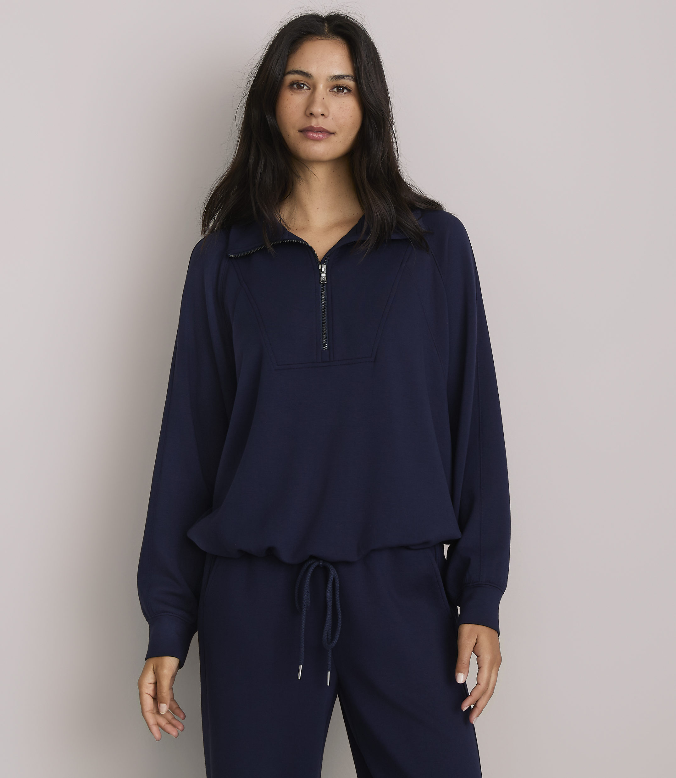 A woman in a navy blue quarter-zip sweatshirt and matching pants stands against a plain gray background.