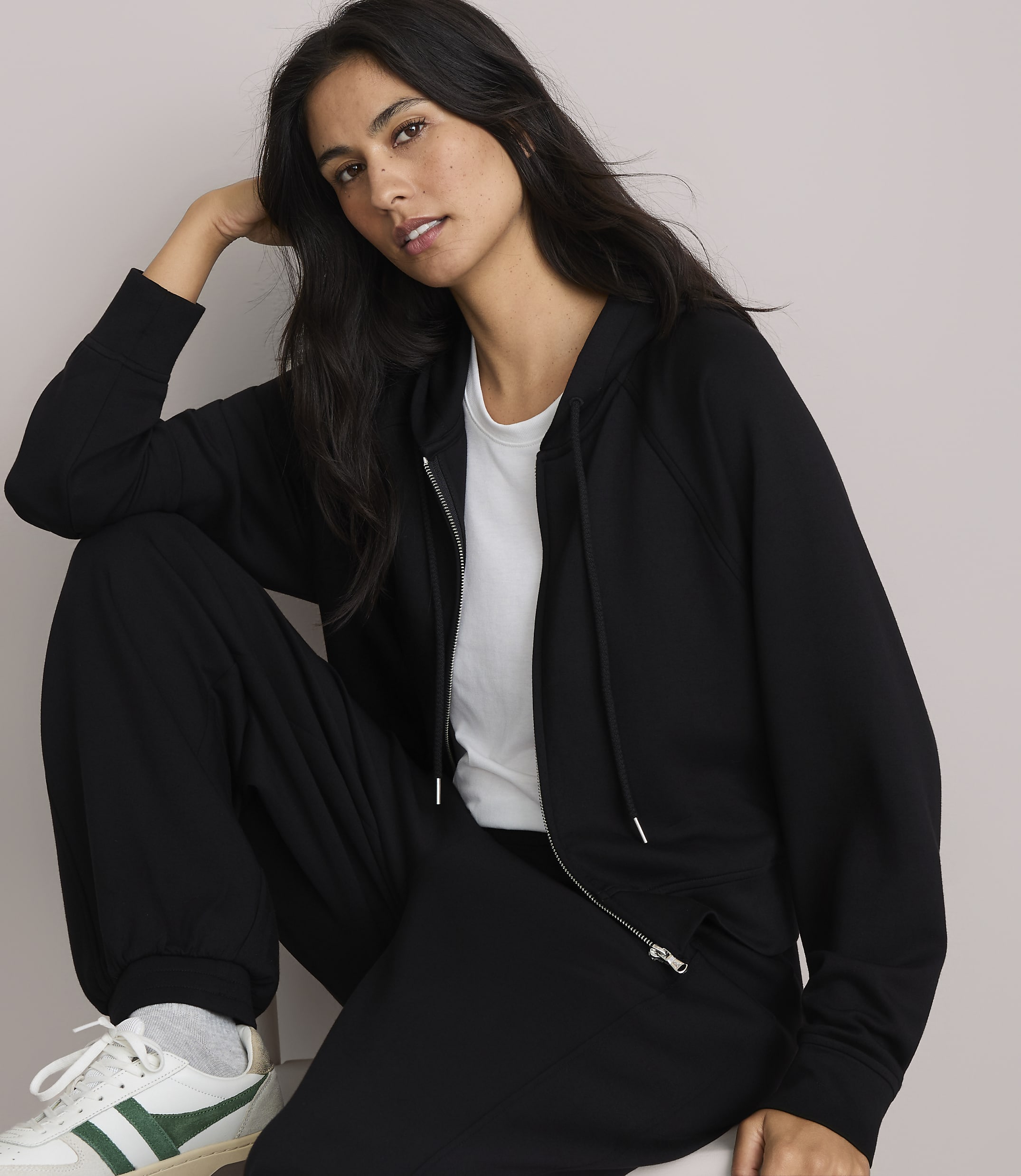 A woman in a black zip-up hoodie and sweatpants poses against a plain, light-colored background.