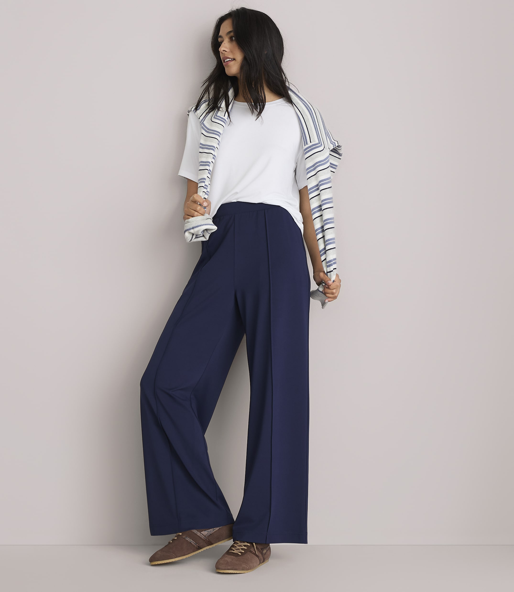 A woman in a white t-shirt and navy wide-leg pants stands against a plain gray background, with a striped sweater draped over her shoulders.